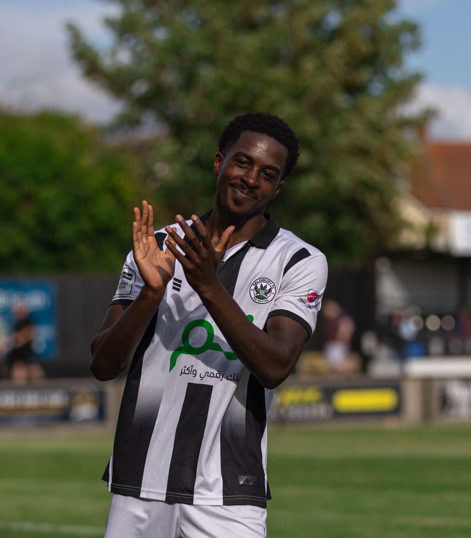 A smiling footballer in black and white kit clapping on a sunny pitch with green trees in the background.