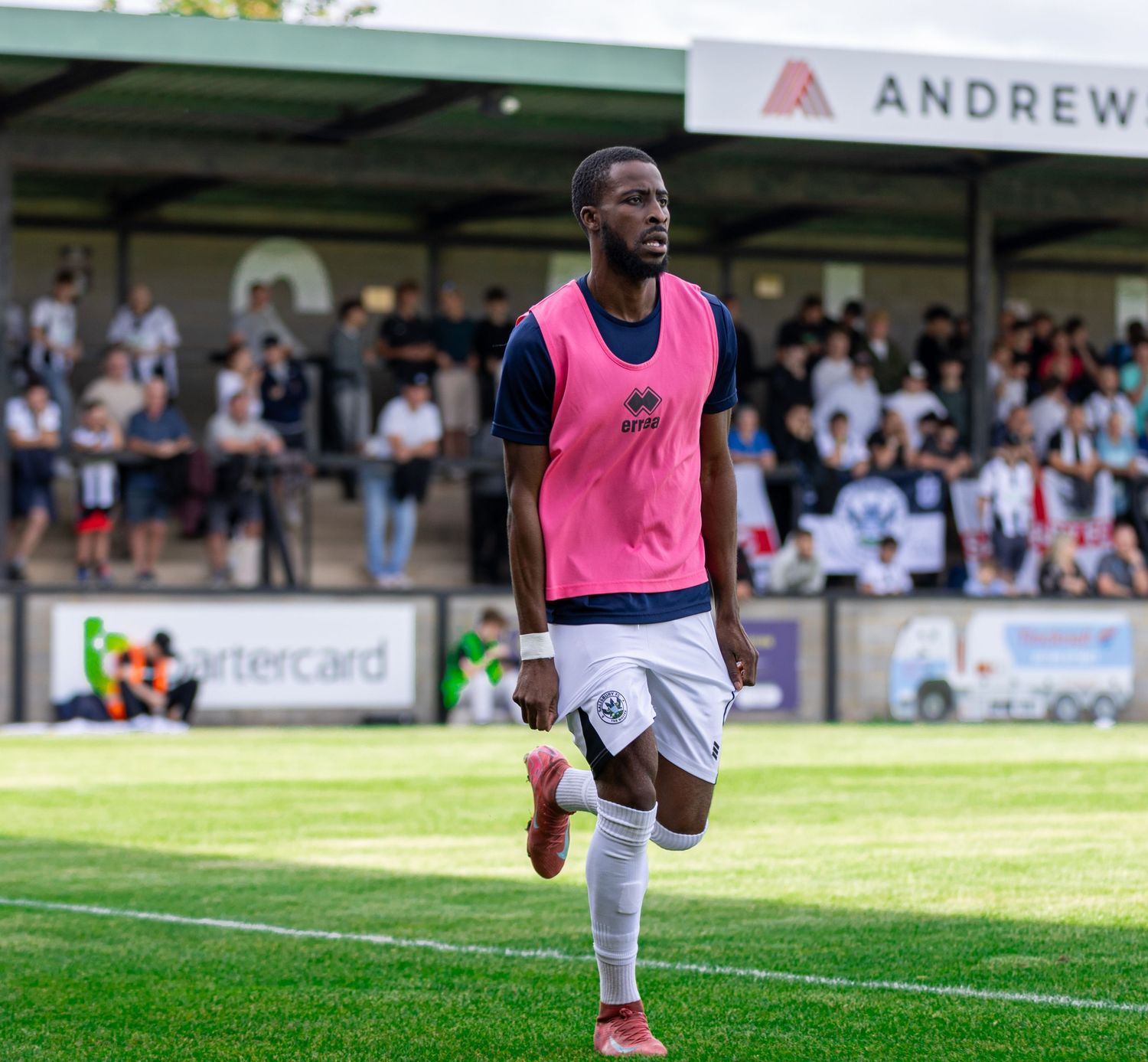 A soccer player in a pink bib and white shorts jogs on the field during a match with spectators in the background.