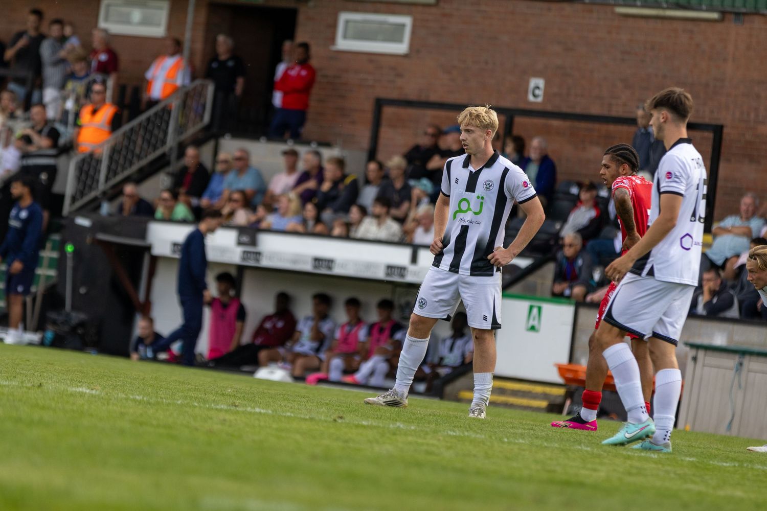 Football players in black and white striped jerseys compete on a green pitch with spectators watching from the stands.