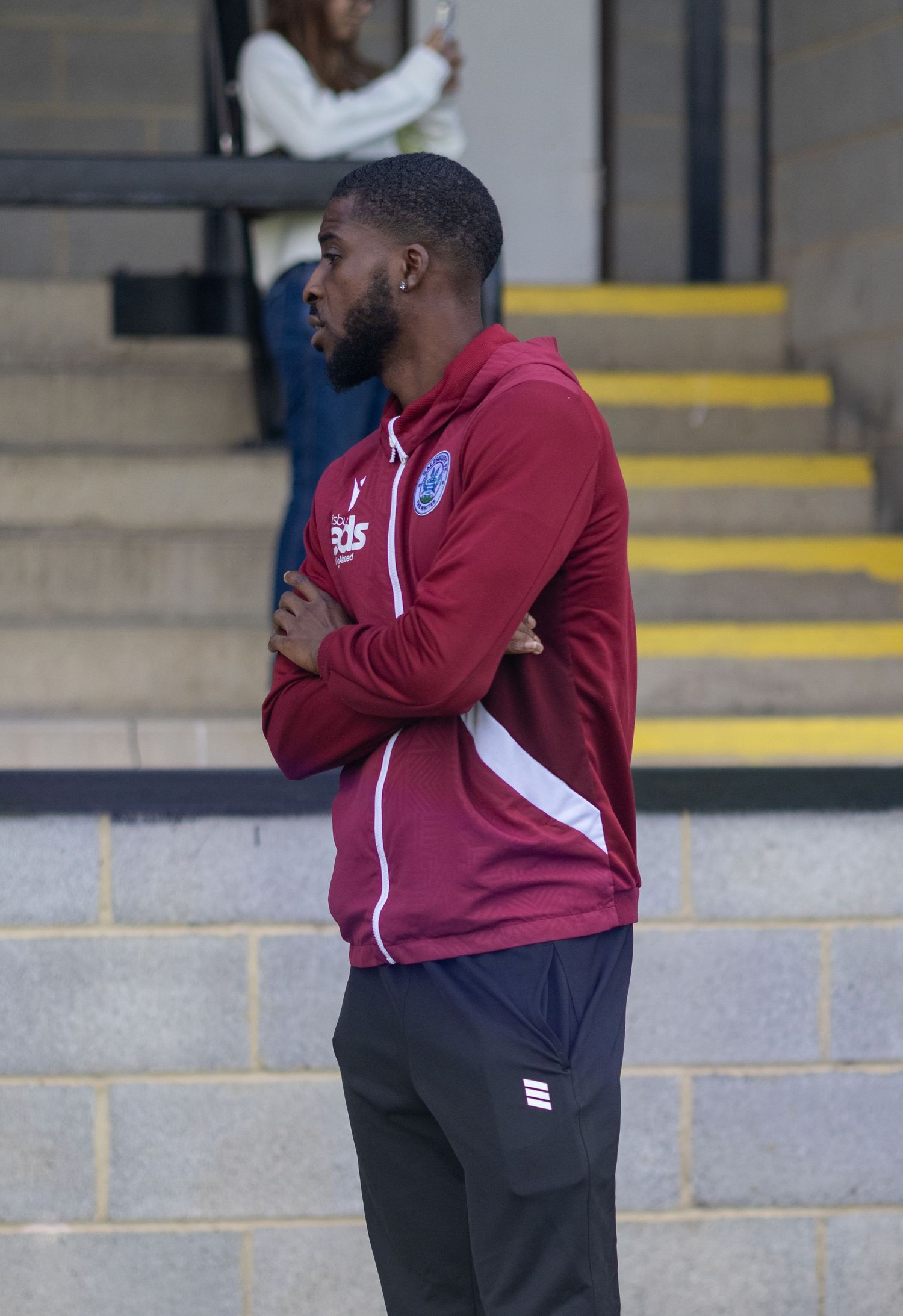 Man in maroon sports jacket with arms crossed standing near stadium steps outdoors.