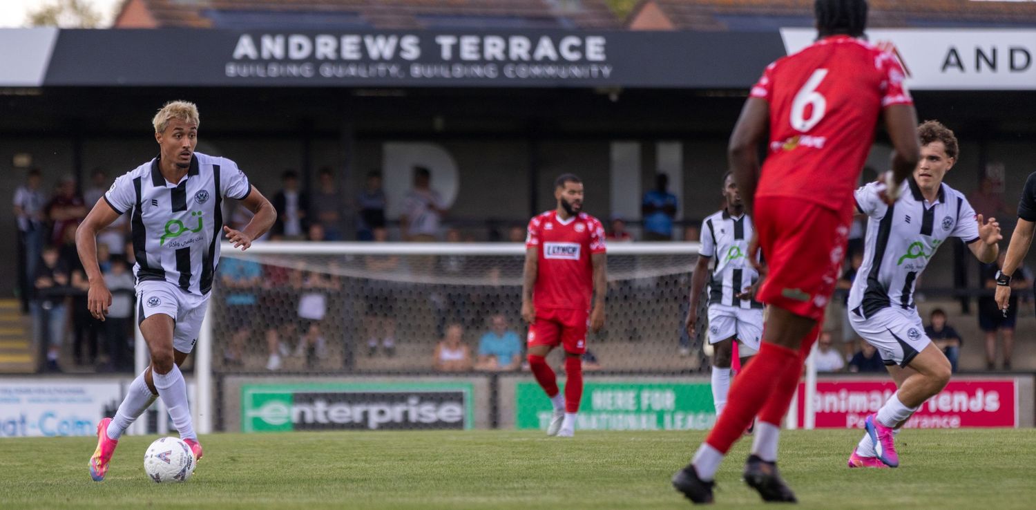 Players in black and white kits control the ball near Andrews Terrace signage during an evening football match.