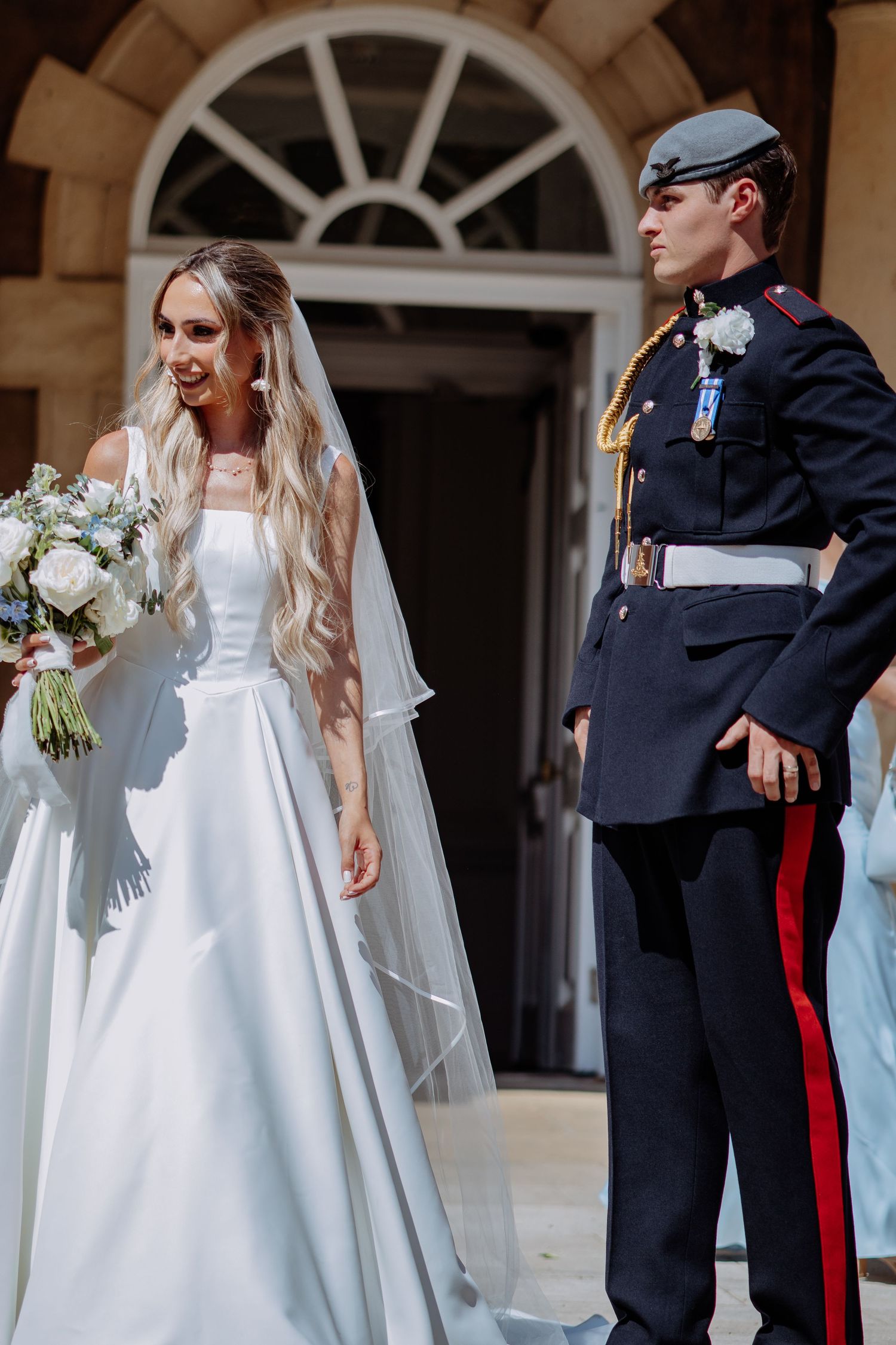 Smiling bride in white satin gown with veil stands beside groom in RAF dress uniform at grand building entrance.