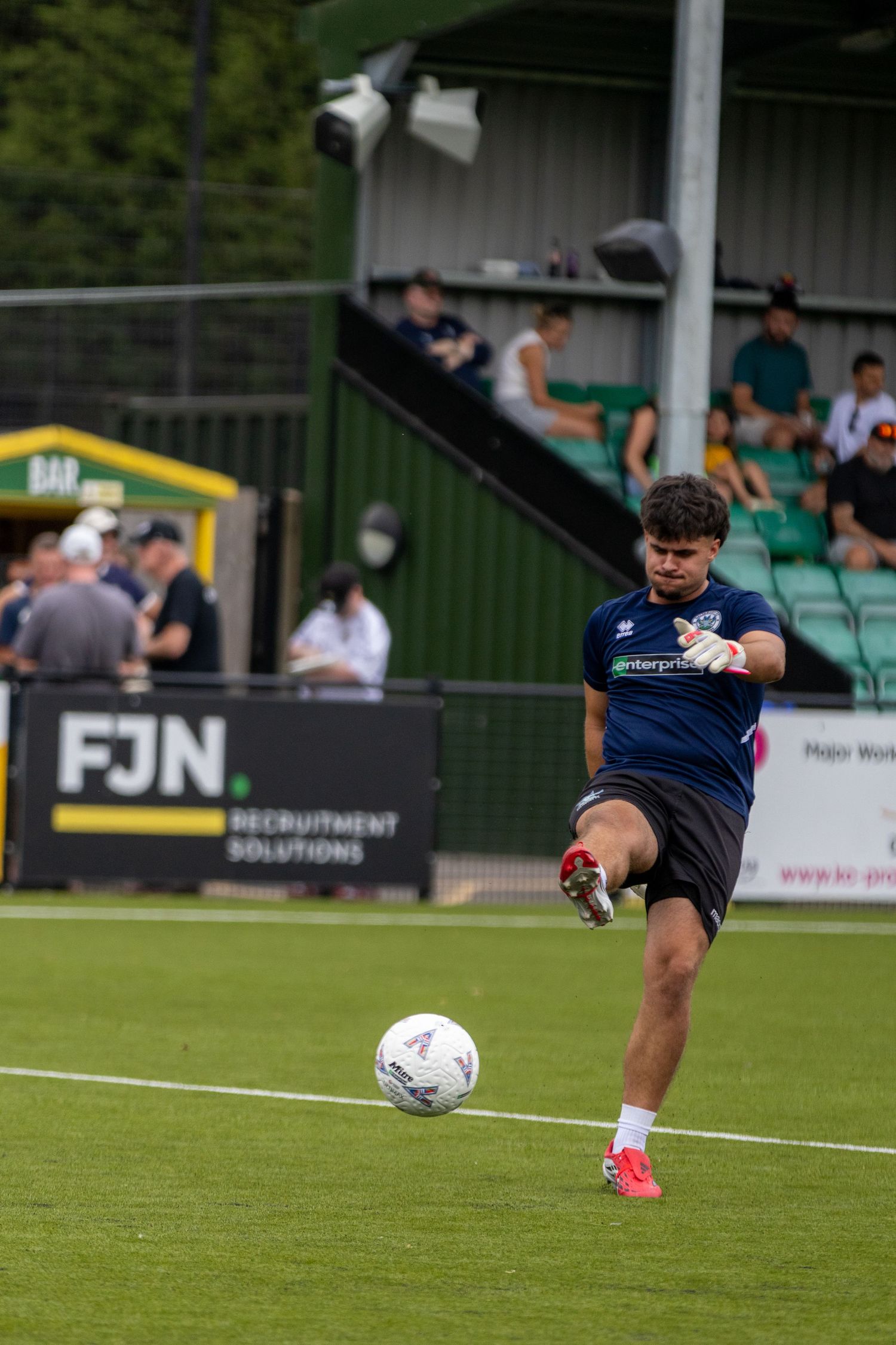 Goalkeeper in navy kit striking ball during warmup drill at non-league football ground with FJN sponsor board visible.