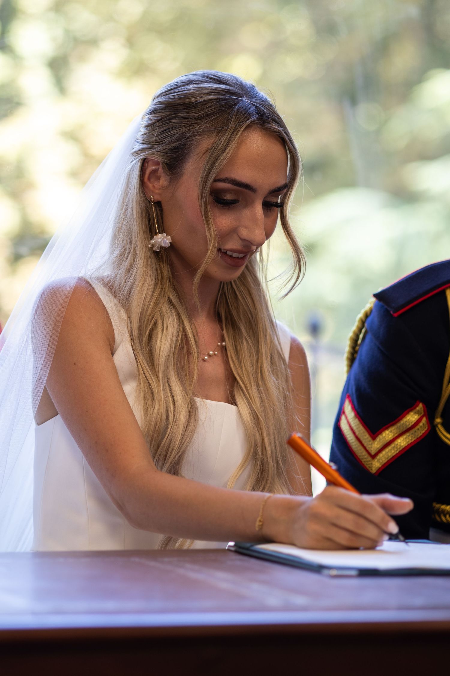 Blonde bride in white gown and veil signing wedding register with orange pen at ceremony.