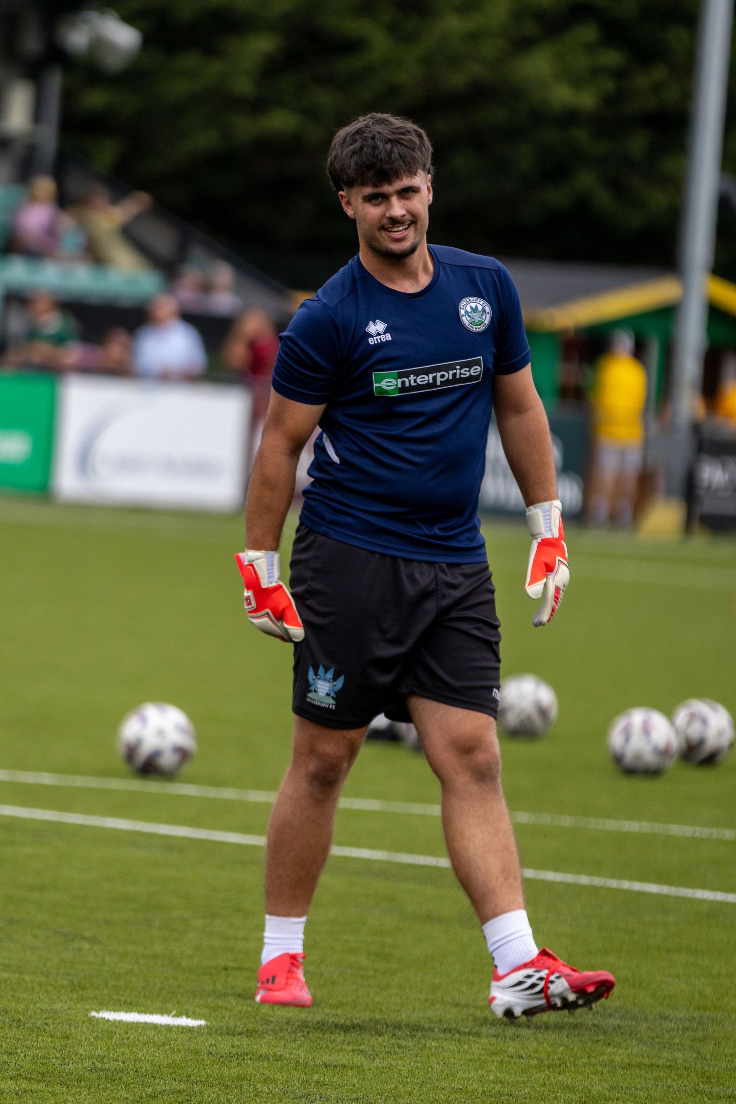 Young smiling goalkeeper wearing navy Enterprise-sponsored football kit and red gloves during pre-match warmup.