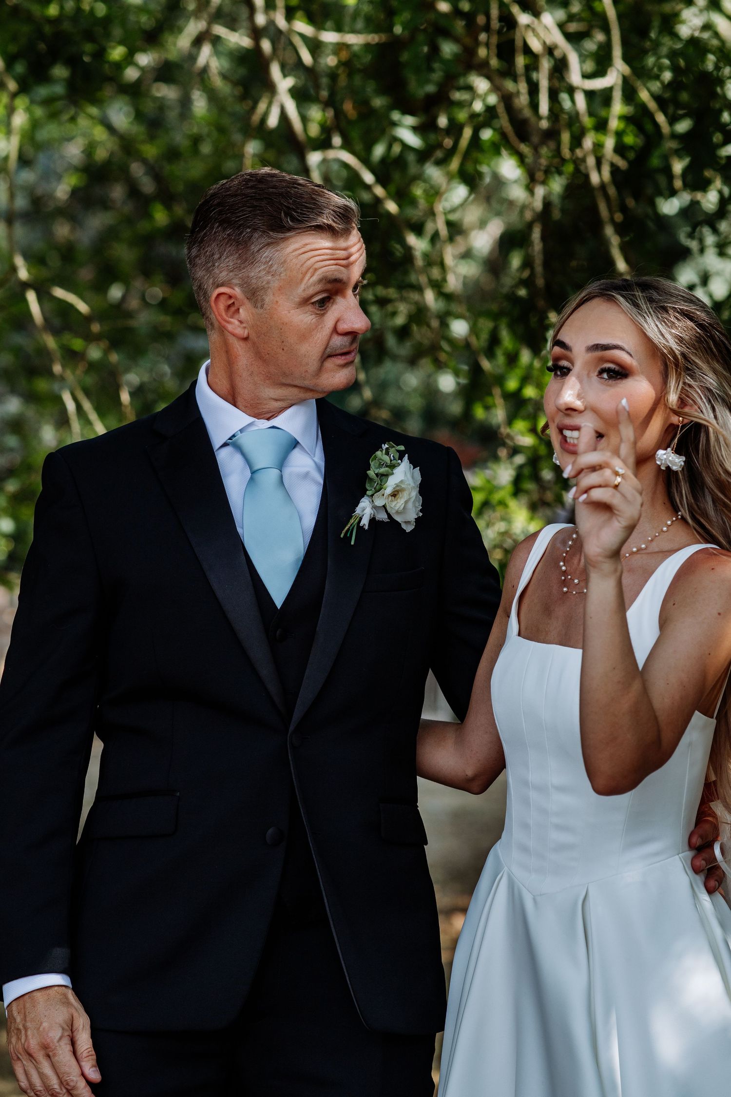 Man in black tuxedo with blue tie and bride in white dress share a joyful outdoor wedding moment.