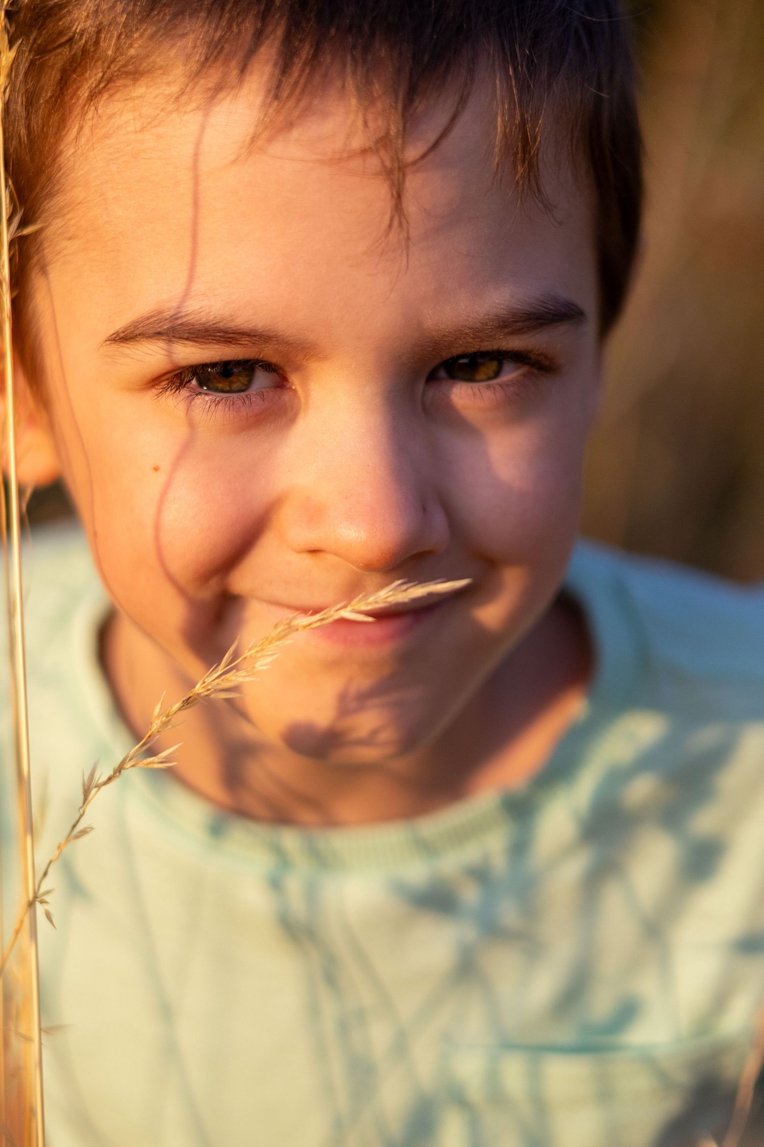 A cheerful young boy smiles warmly while holding a grass stem in glowing sunset light.
