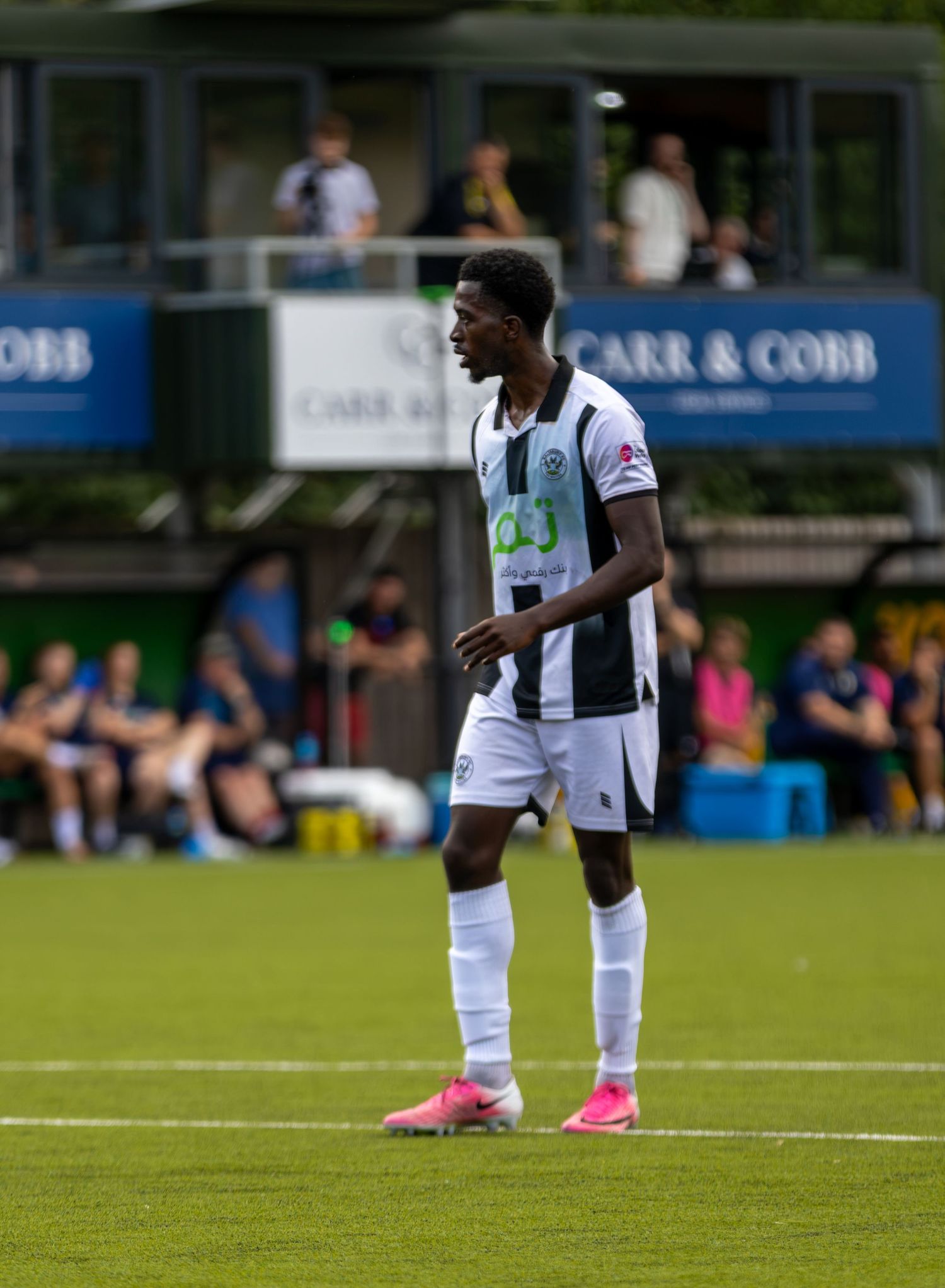 A Black footballer in white and black striped kit stands on the pitch during a sunny football match.