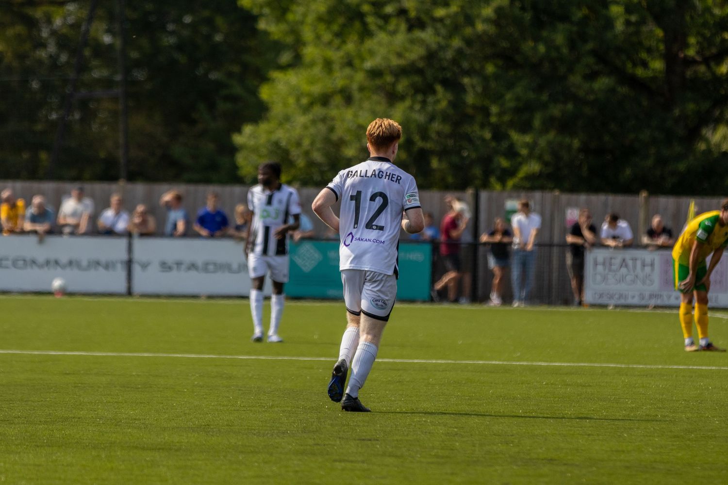 A footballer wearing number 12 in black and white striped kit runs across a green pitch during a sunny match.