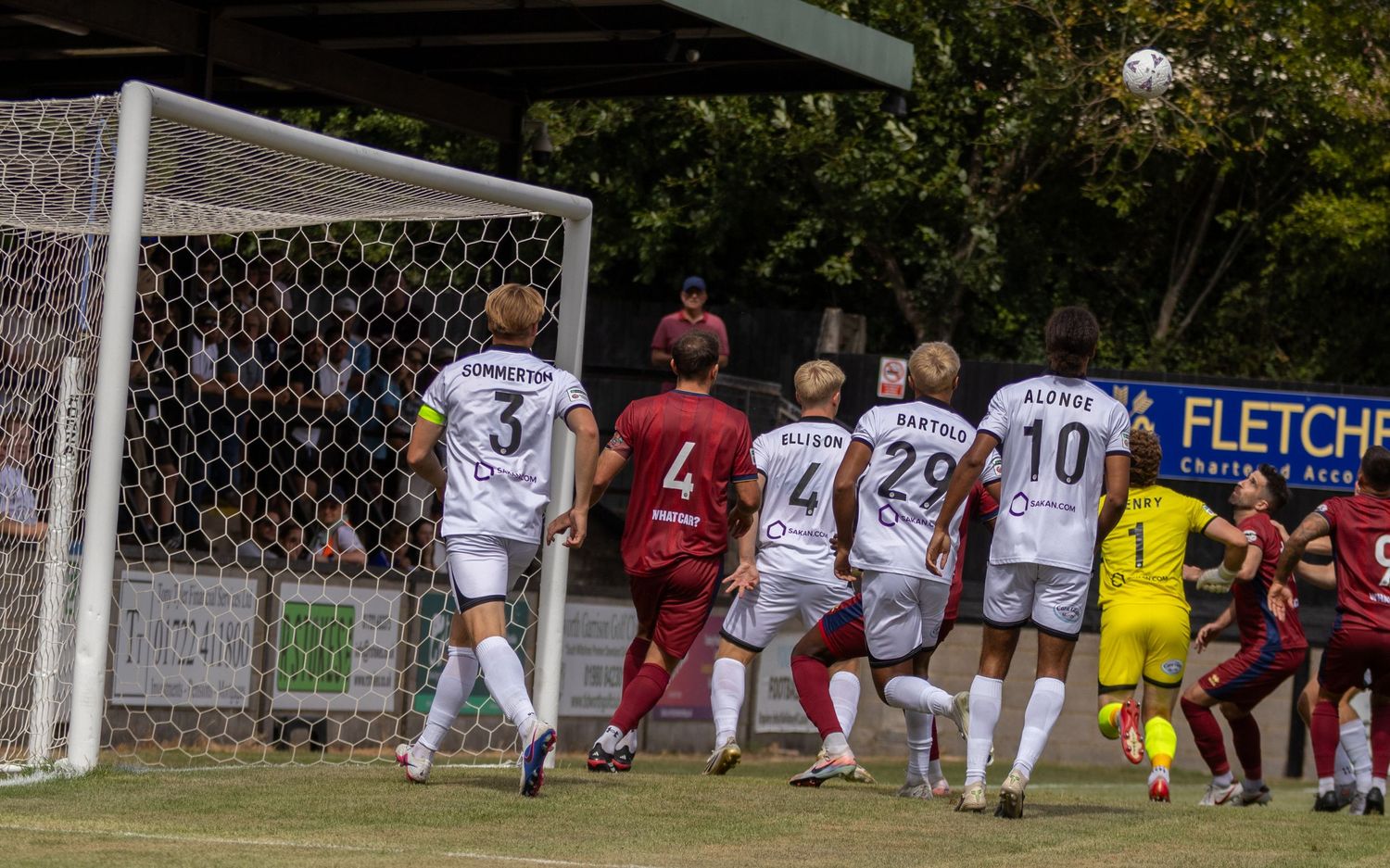White-kit players including number 29 BARTOLO defend goalmouth during corner kick in football match.