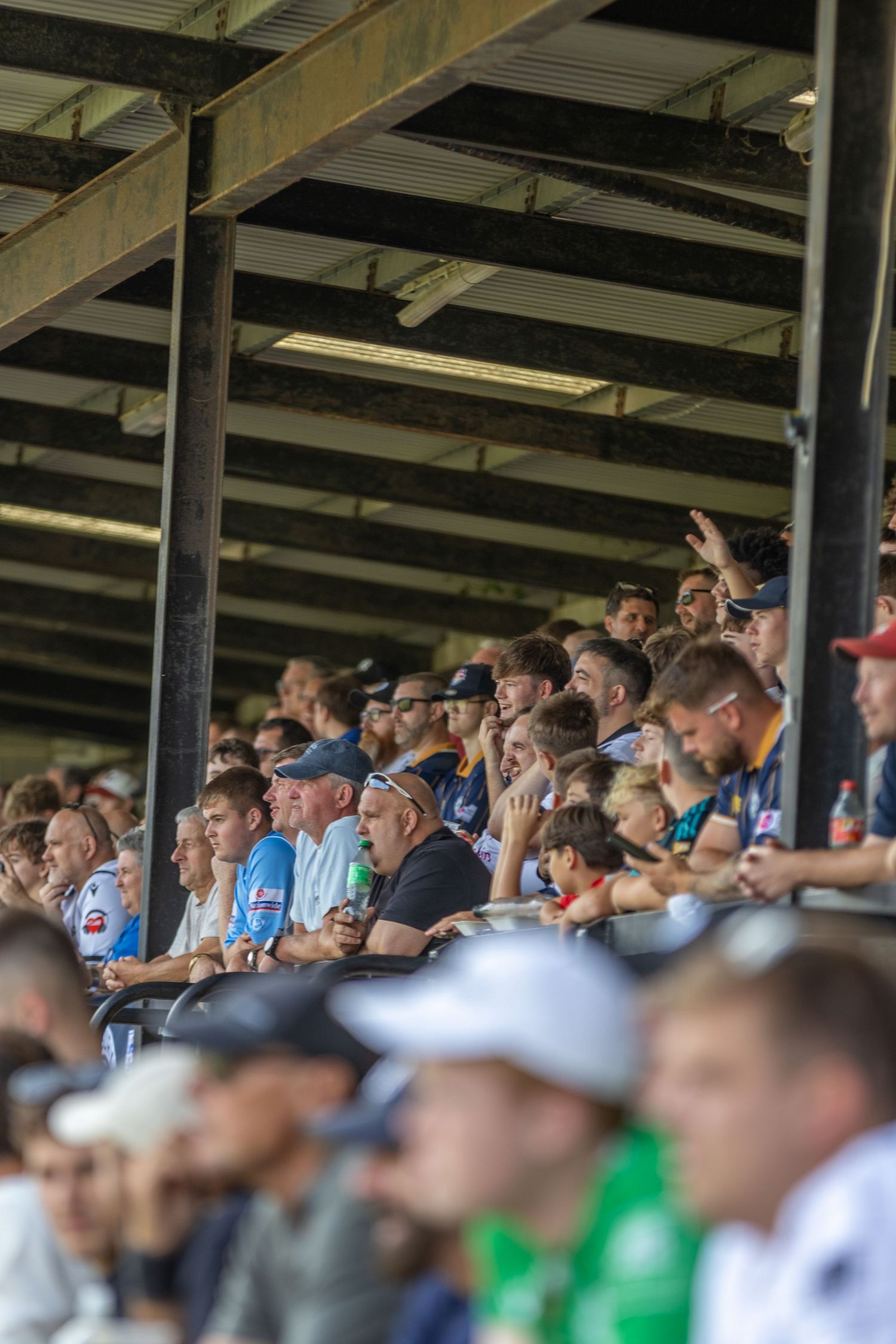 Crowds of football fans watch a match from a covered stadium stand on a sunny day.