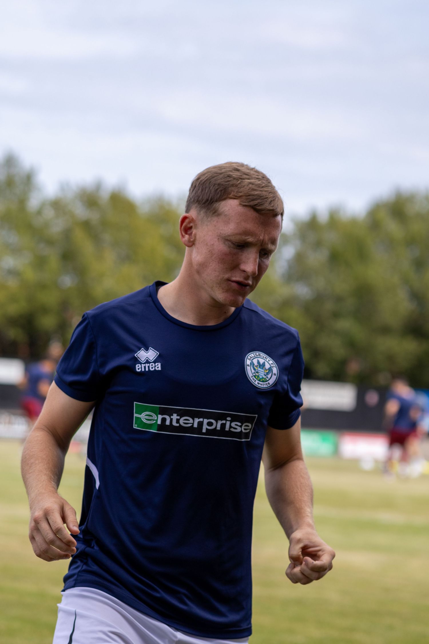 Male footballer in navy Enterprise-sponsored Errea shirt jogging during pre-match warmup on football pitch.