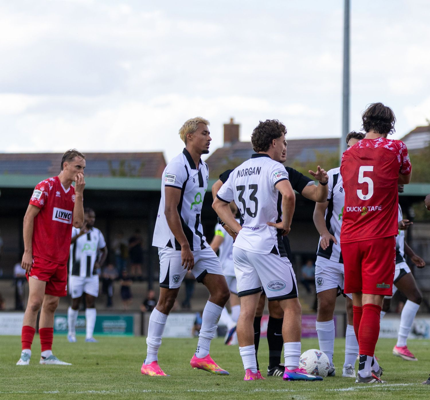 Players in red and white kits gather around the ball during a non-league football match, including Morgan 33.