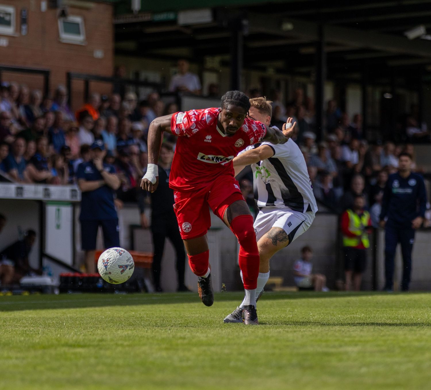 Red-kitted footballer battles for ball against white-shirted defender in front of packed stadium crowd.