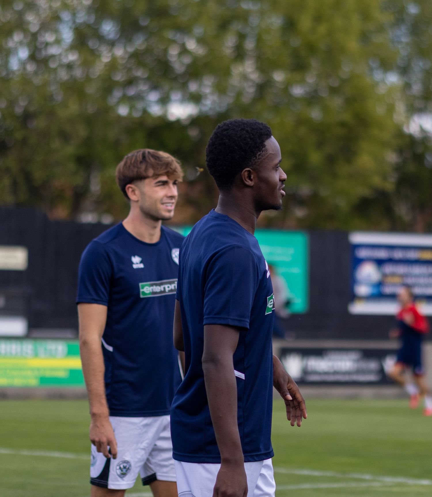 Two male football players in navy Errea kits smile and chat during a pre-match warmup on a grass pitch.