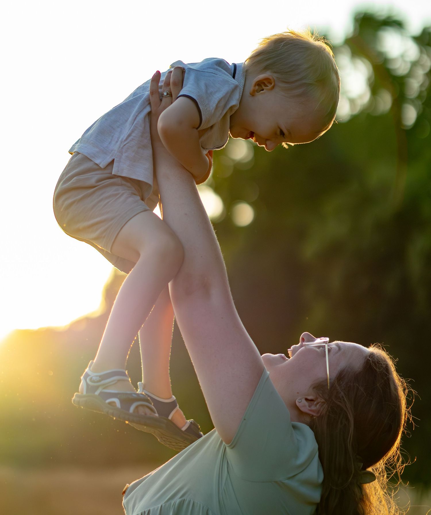 A happy mother raises her young child overhead against a warm glowing sunset background.