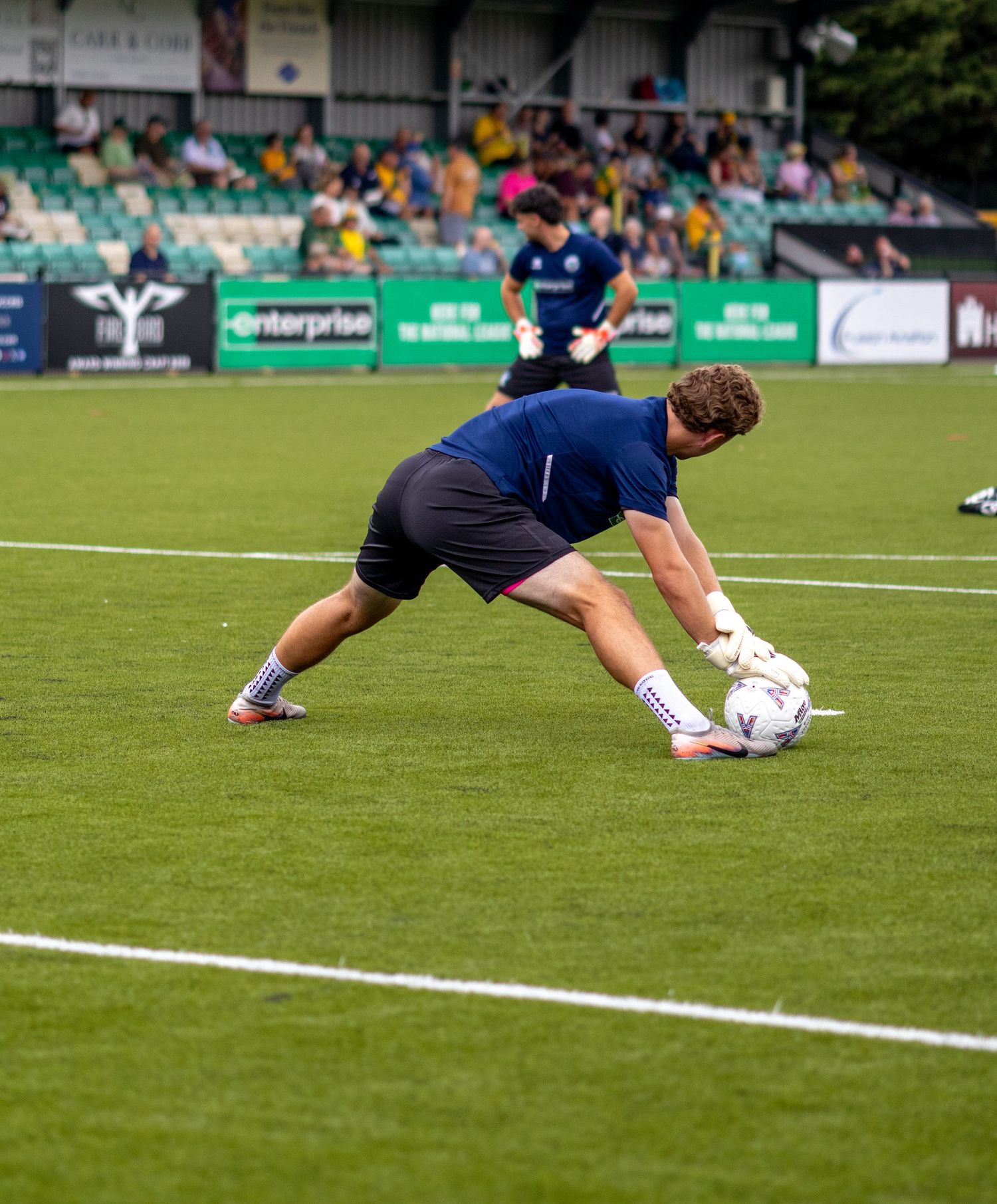 Goalkeeper in blue kit diving to collect football during warm-up on green pitch.