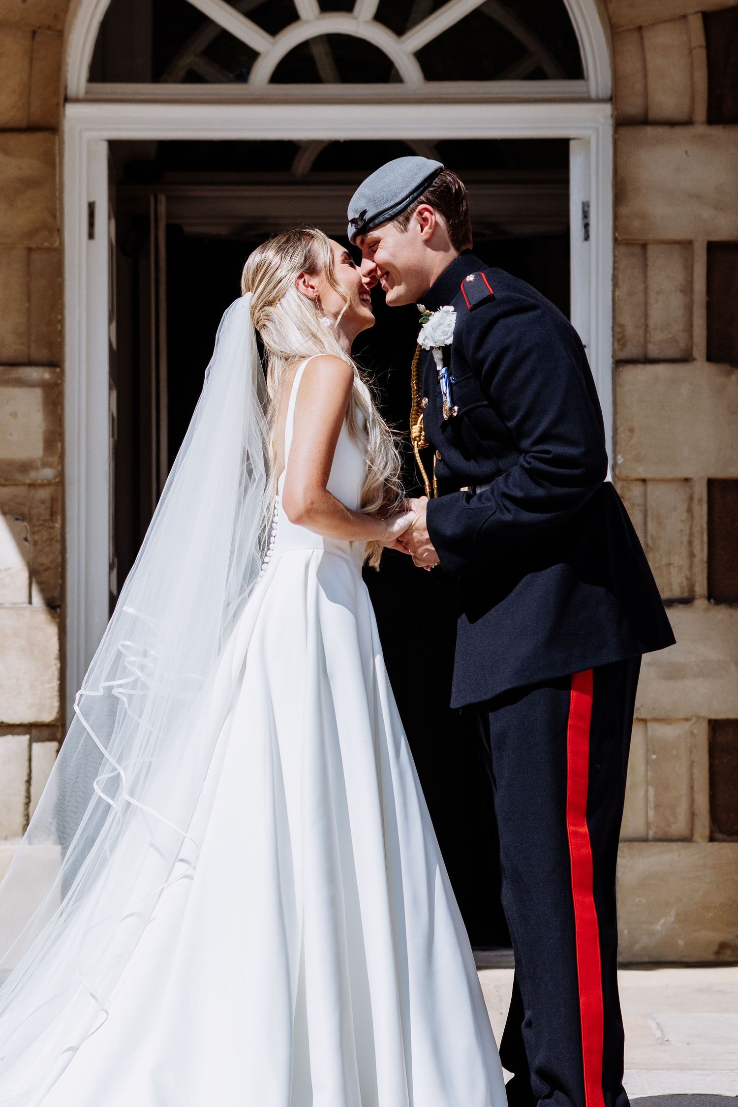 Bride in white ball gown and veil leans in close with groom in military uniform holding hands outdoors.