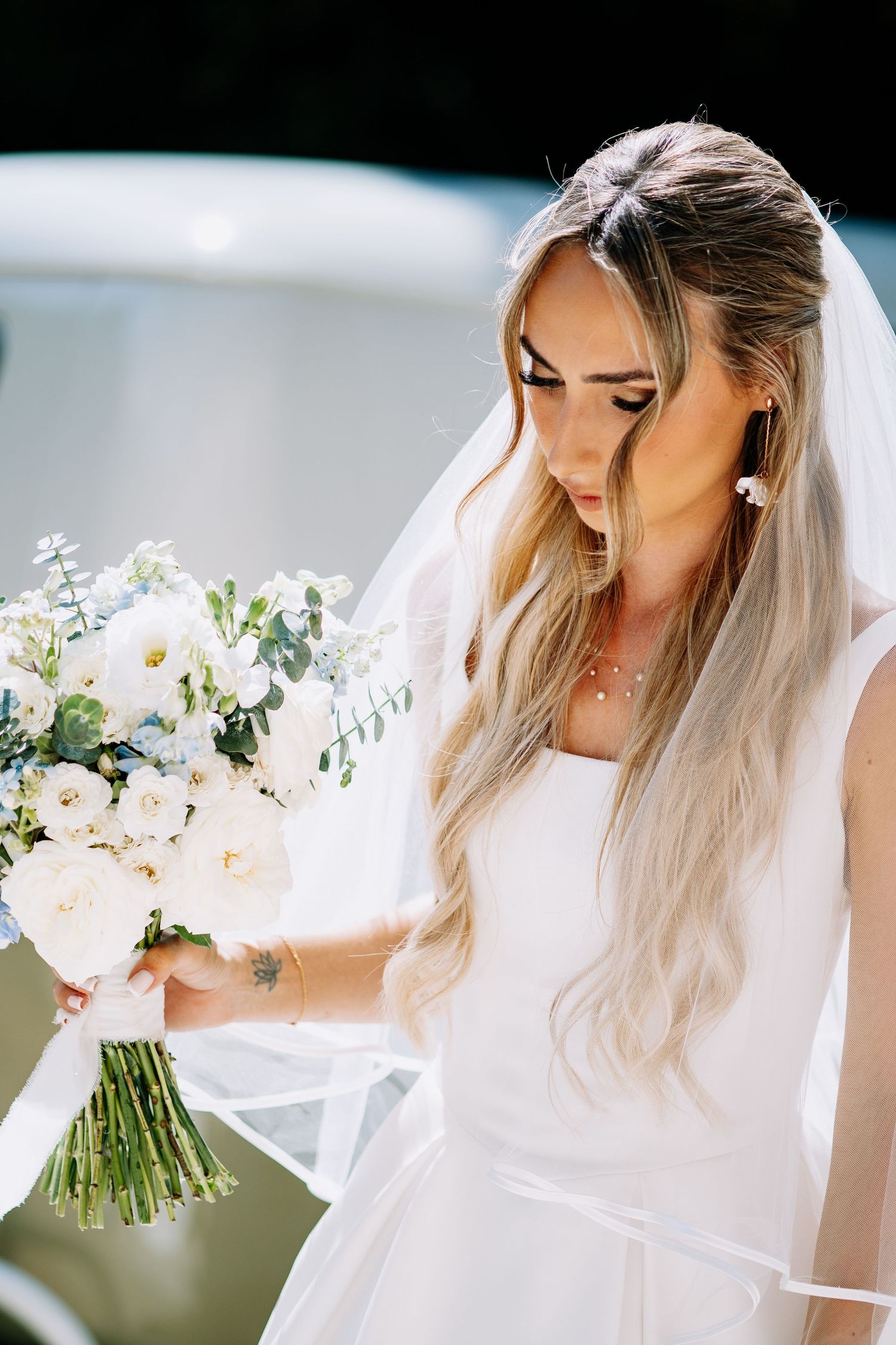 Close-up of bride in white gown with veil admiring her white rose and eucalyptus bouquet.