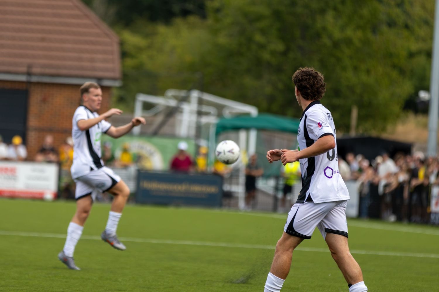 Two white-kitted footballers compete for the ball near the goal during an outdoor football match.