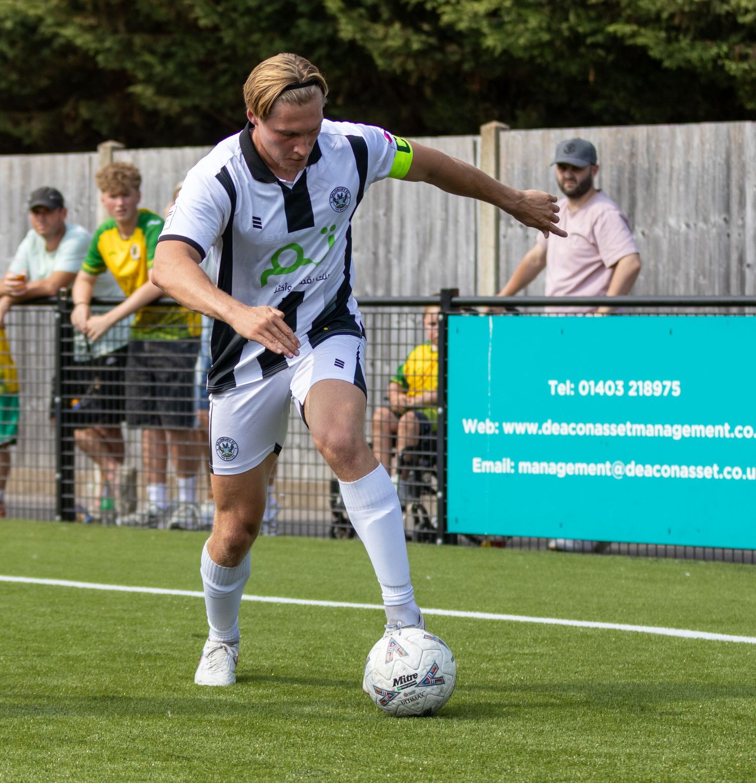 A footballer in white and black kit controls the ball during an outdoor match on a green pitch.