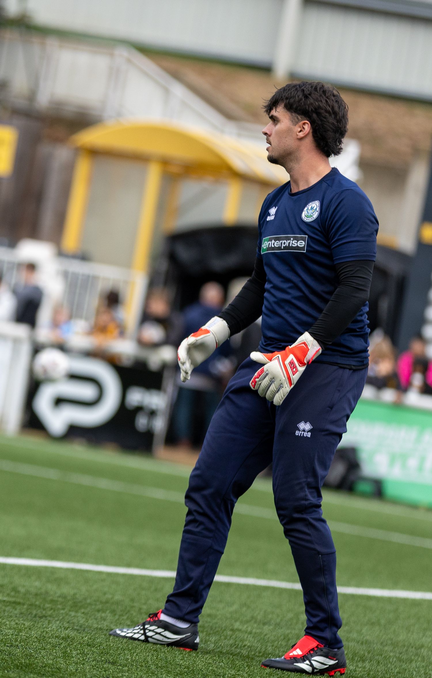 A goalkeeper in navy blue kit and red gloves stands alert on a football pitch during a match.
