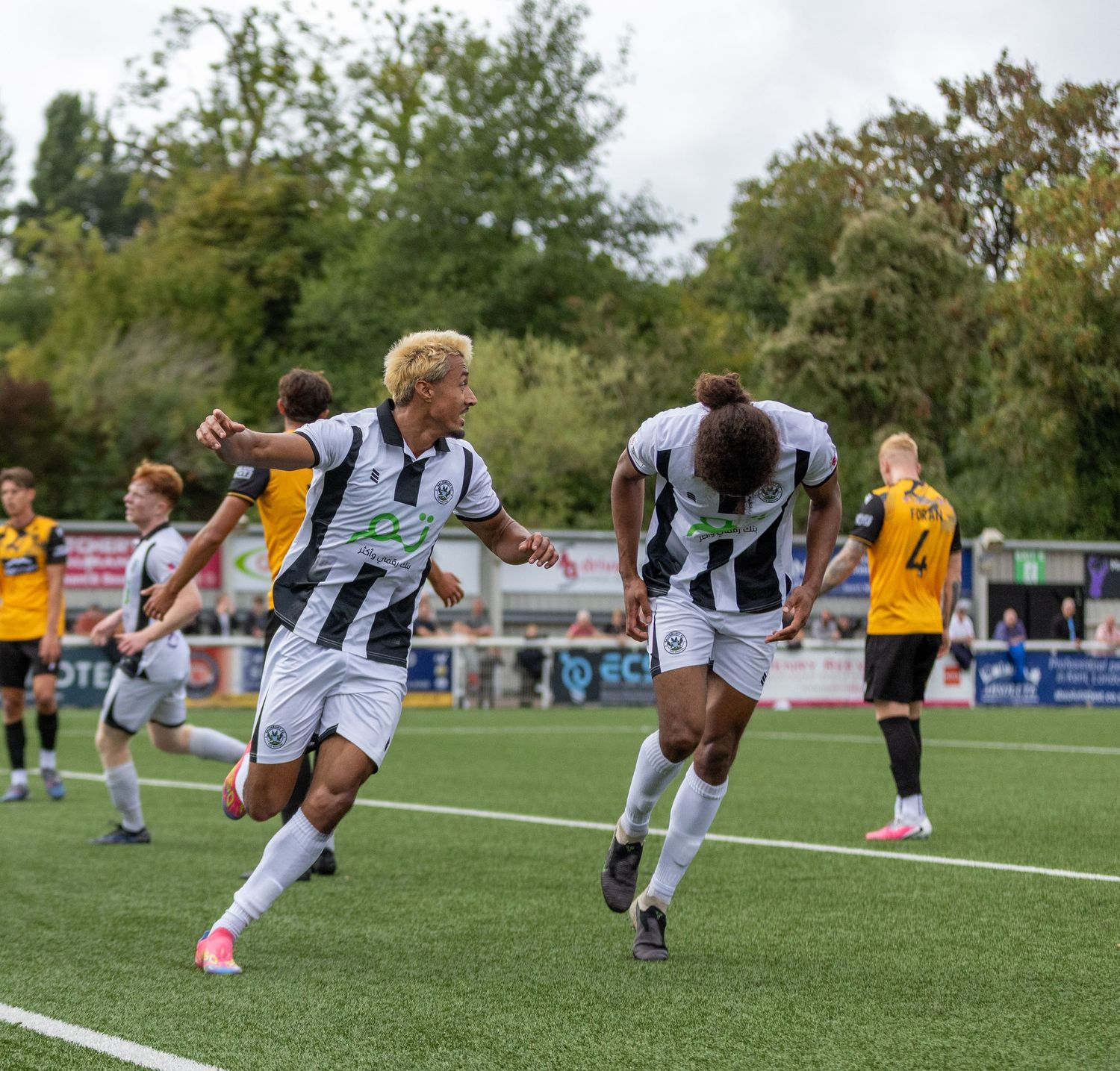 Two soccer players in white uniforms celebrate a goal during a match on a green field with fans in the background.