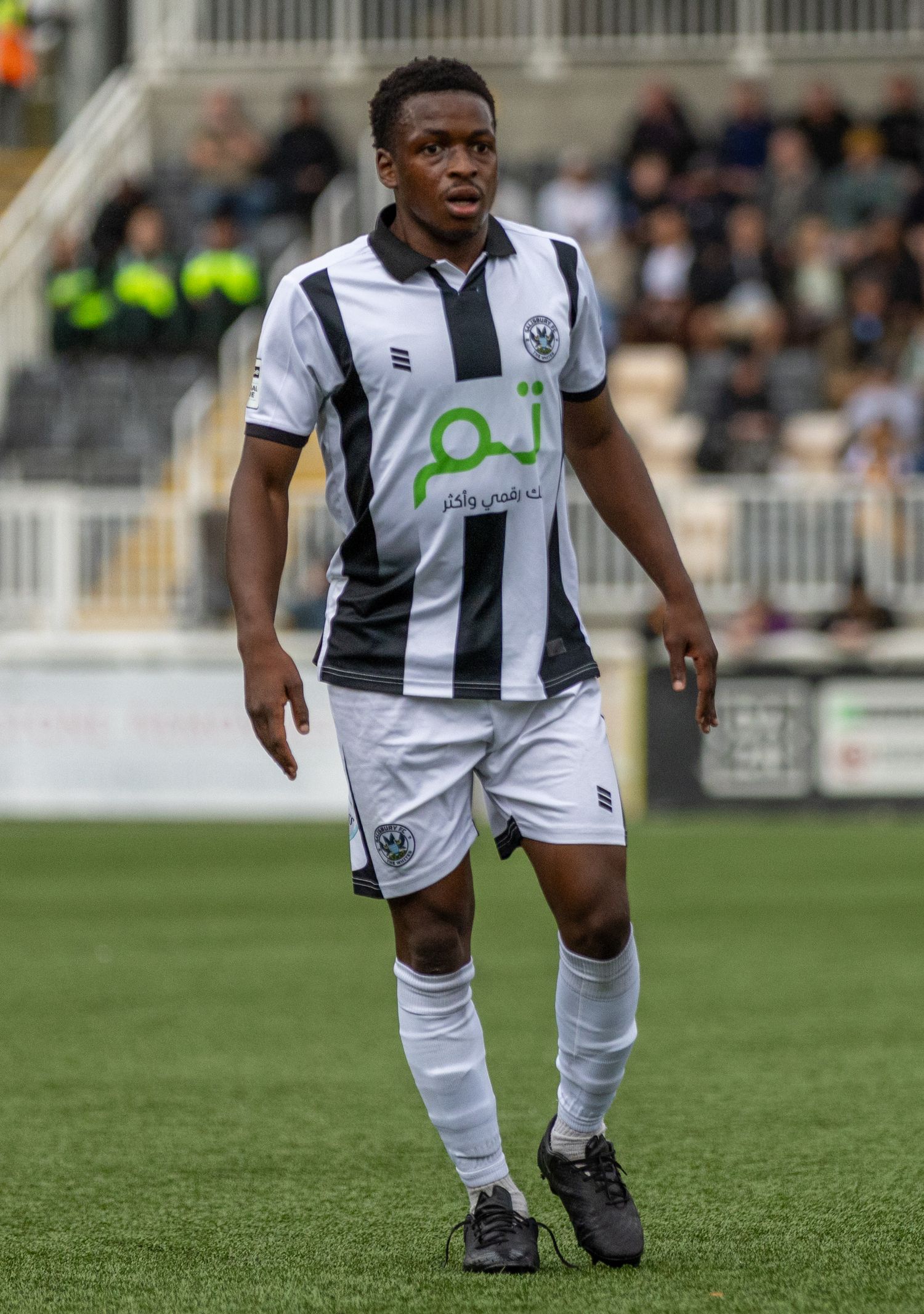 Soccer player in black and white striped kit with Arabic sponsor logo stands on green pitch during match.