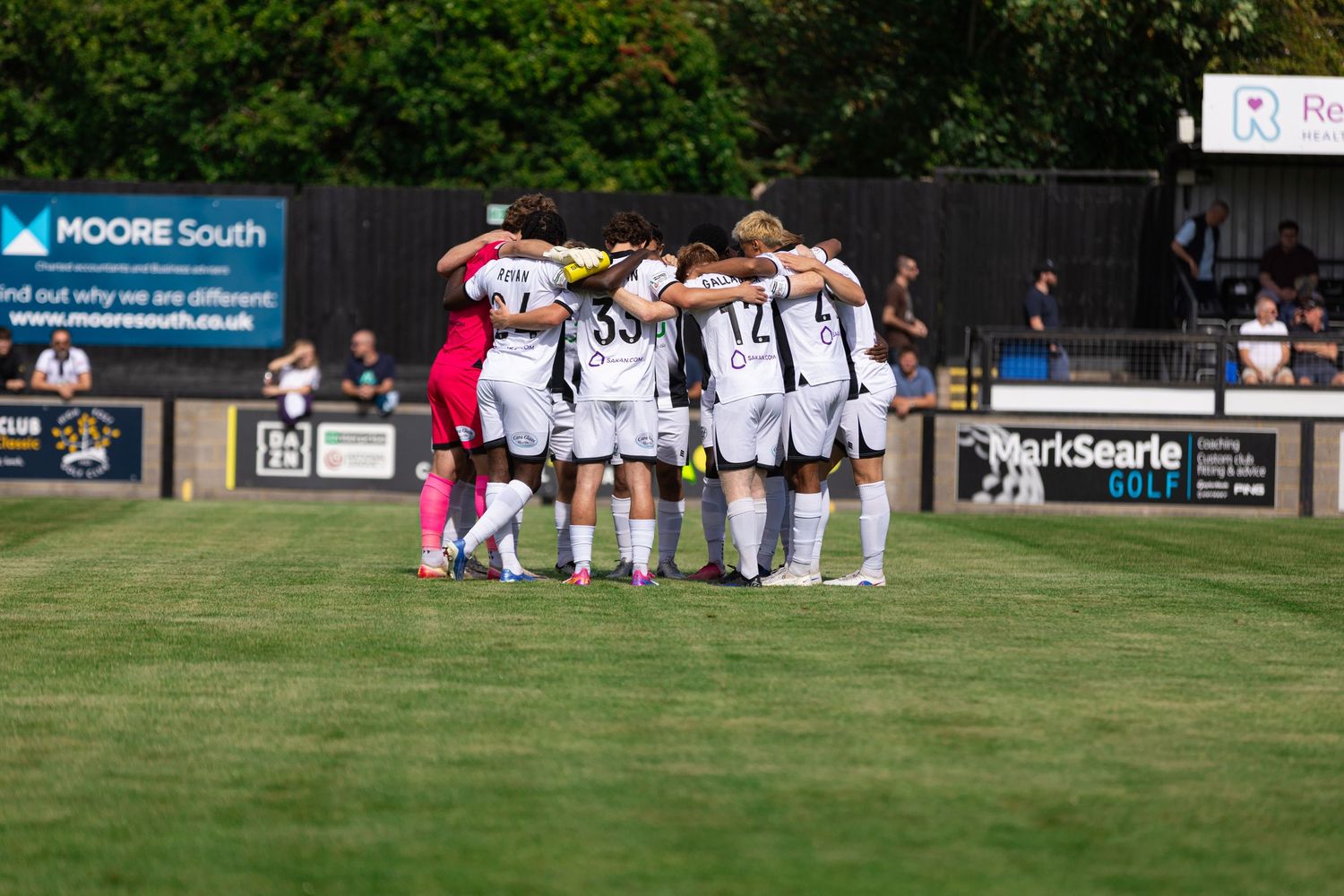 Football team huddles together on a sunny pitch before a match, wearing black and white kits.