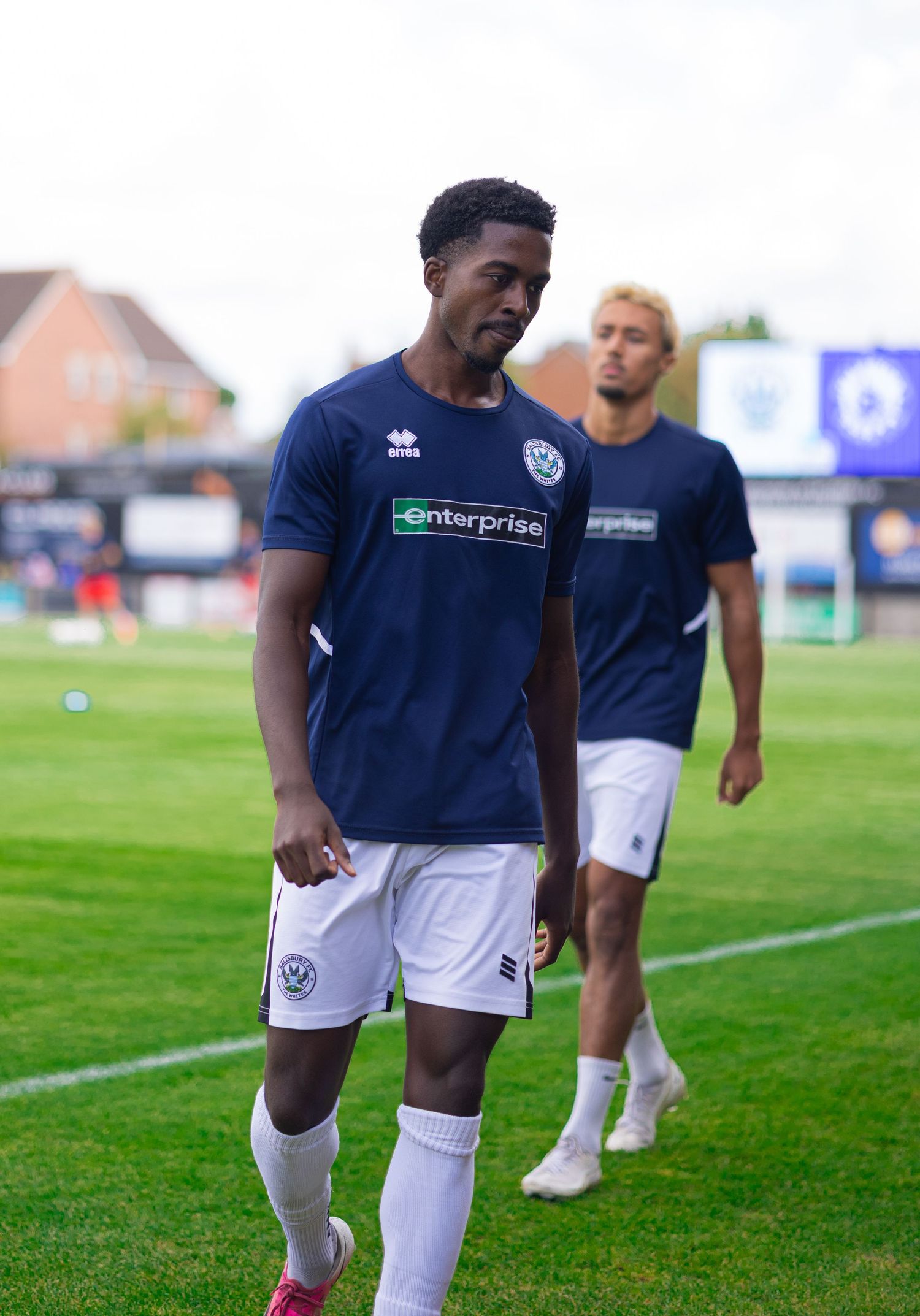 Two football players in navy Enterprise-sponsored jerseys walk on a green pitch during a match.