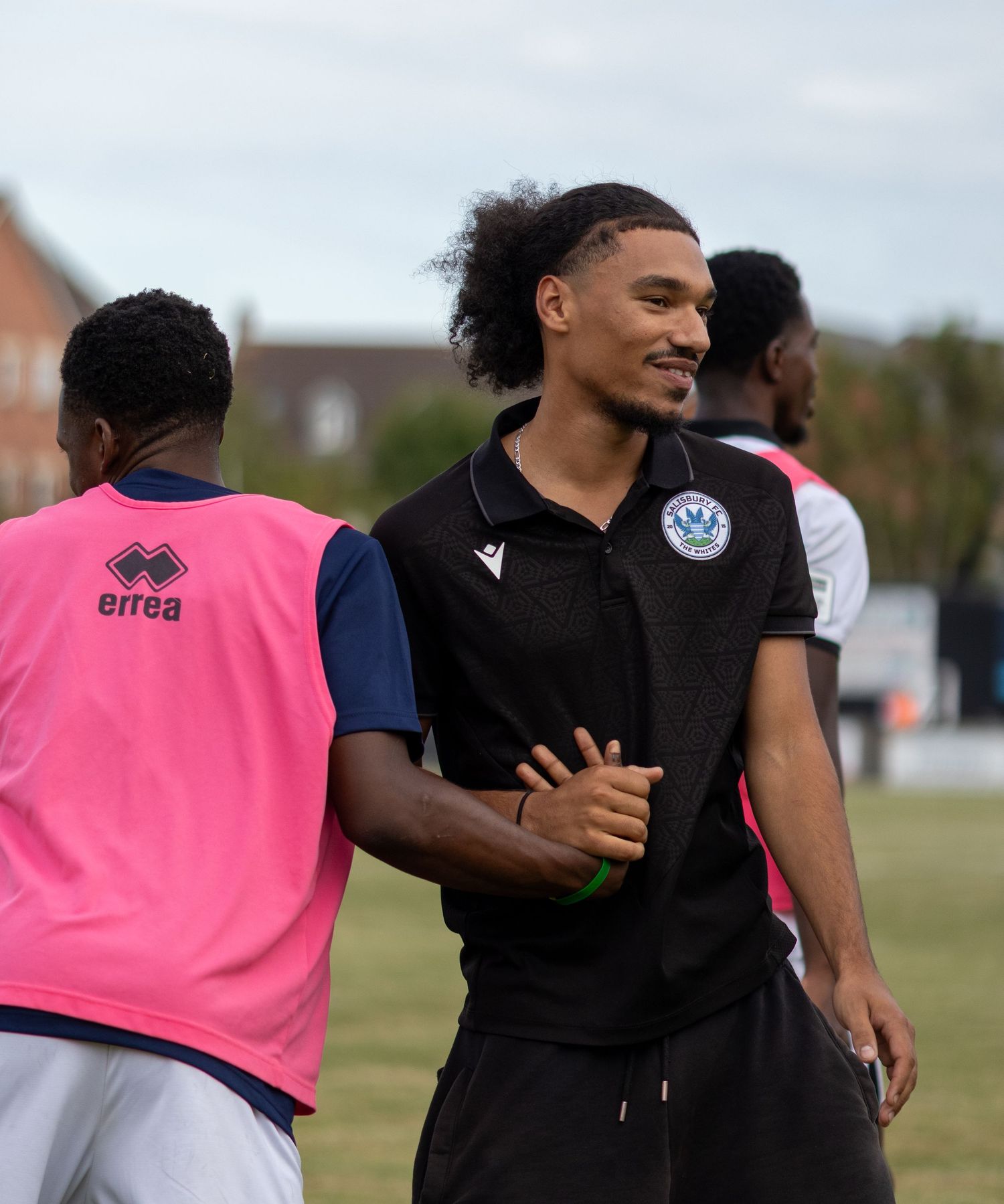 Smiling footballer in black polo shirt greets teammate in pink training bib on the pitch.