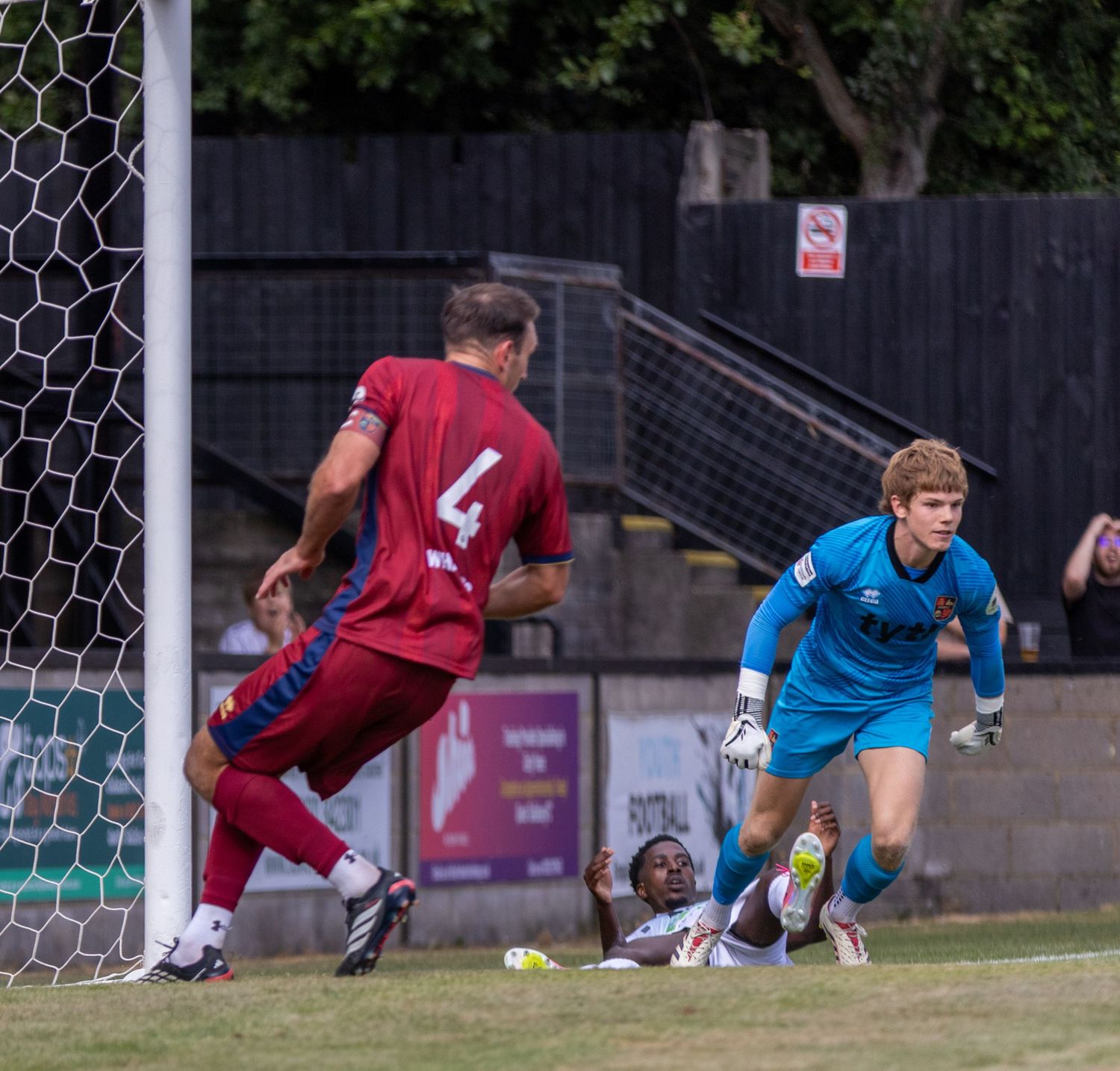 A goalkeeper dives as an opposing striker scores a goal during a non-league football match near the net.
