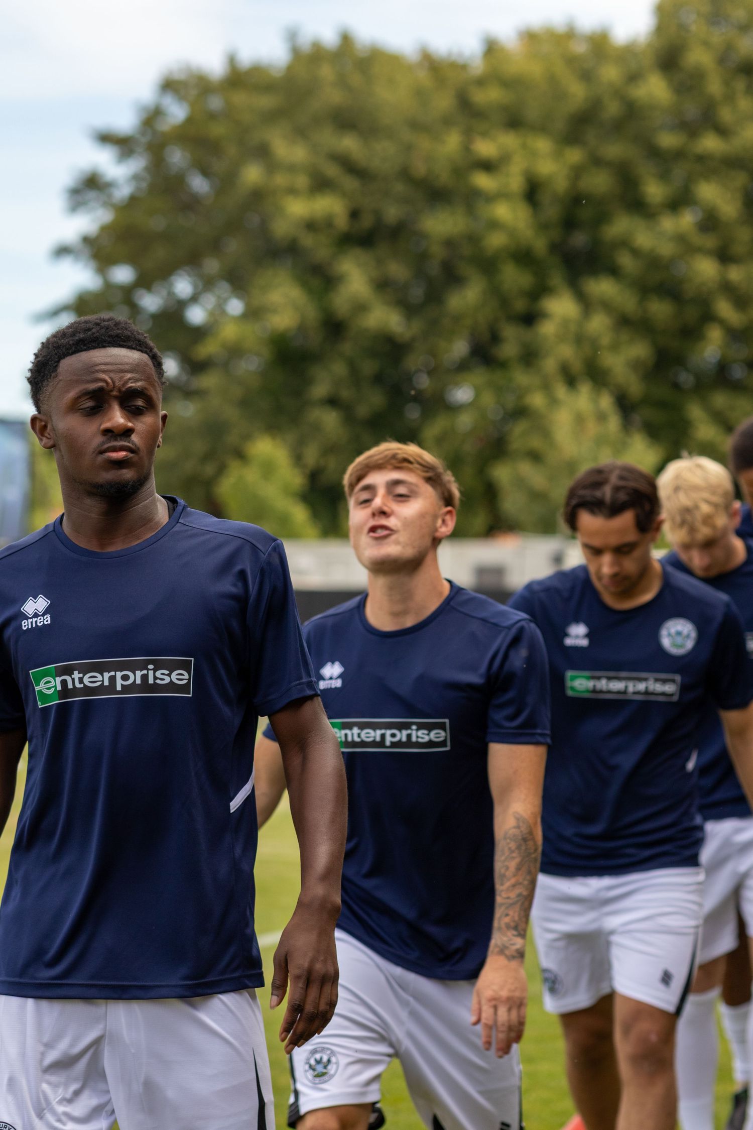 Amesbury FC players in navy Enterprise-sponsored Errea kits walk across the pitch before a football match.