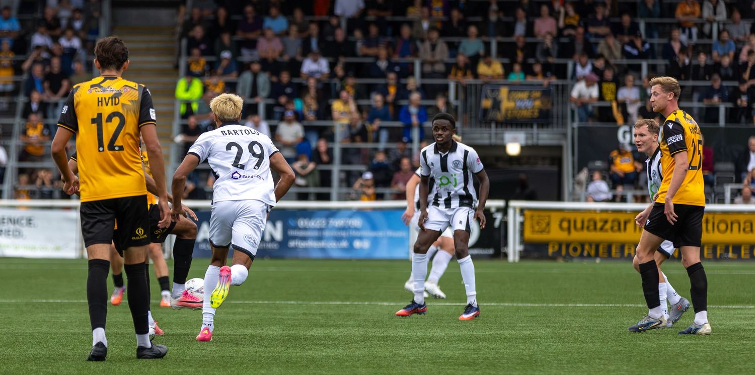 Football players compete on a pitch during a match, with yellow and black kit players facing white and black striped opponents.