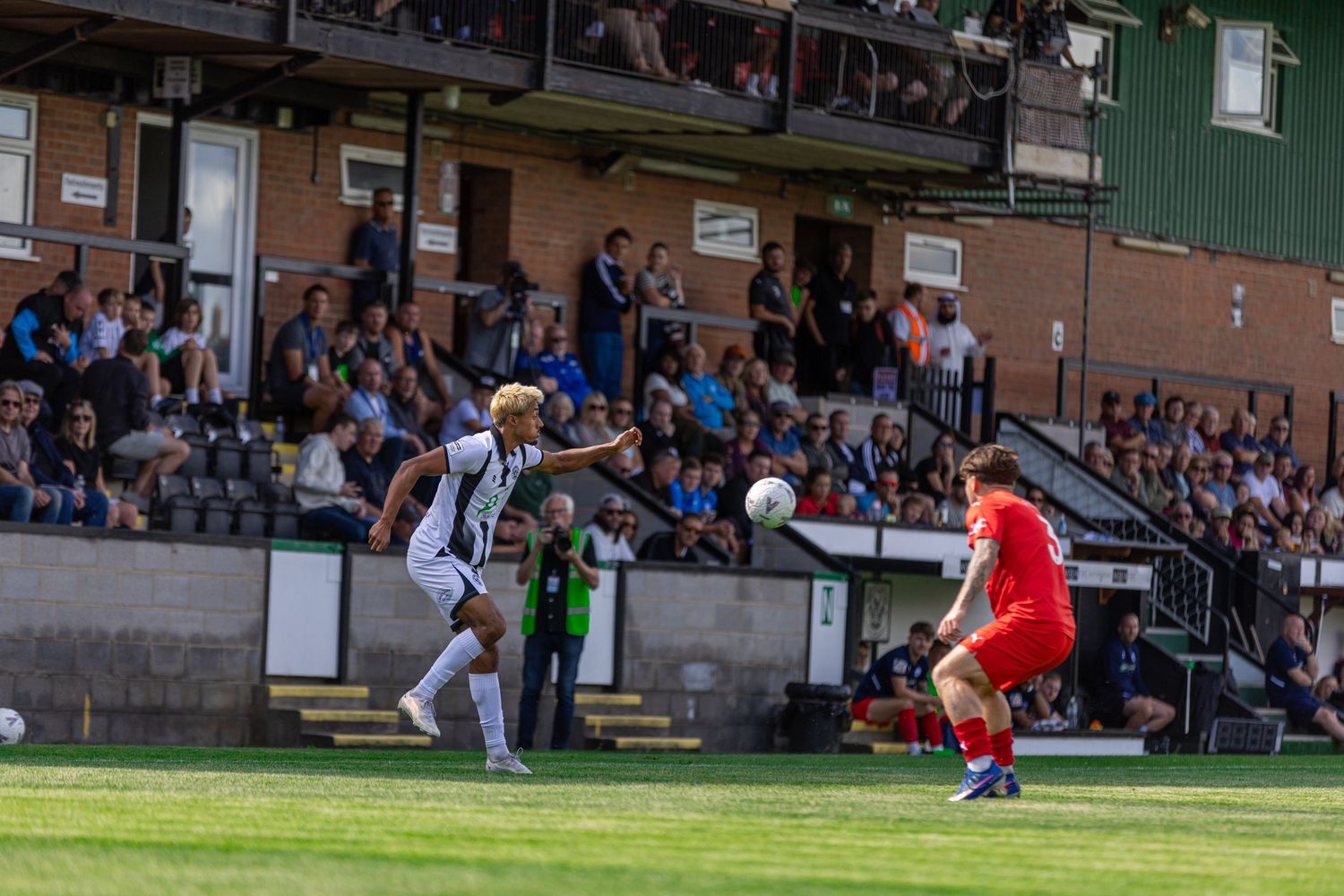 Two soccer players compete on a green pitch during a match with spectators watching from the stands behind them.