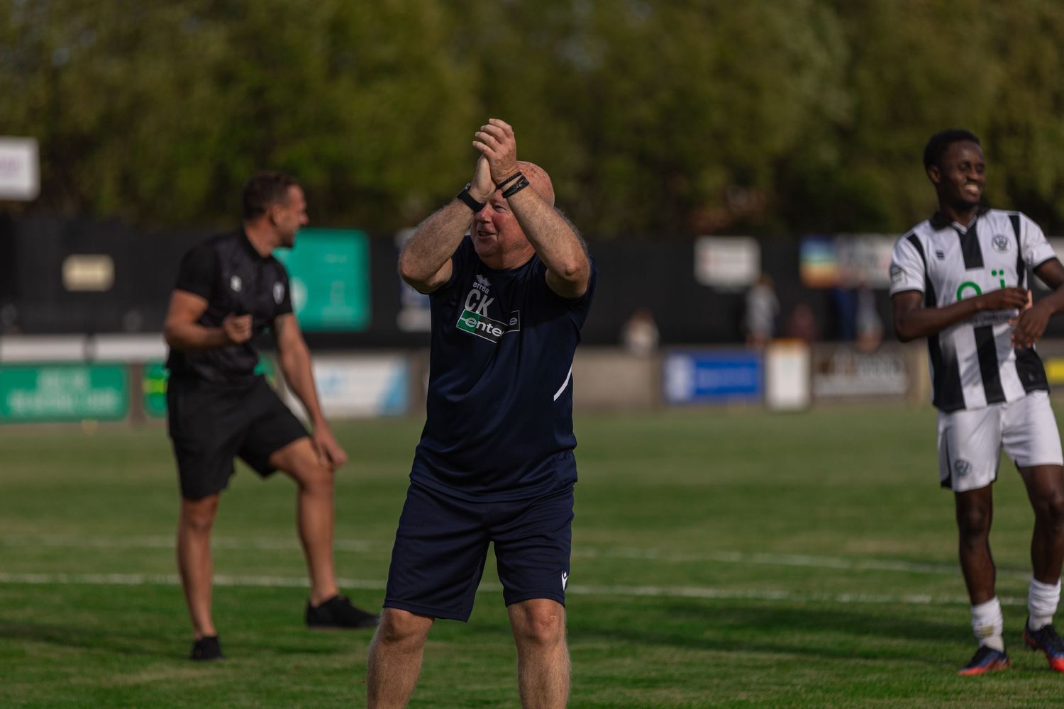 A football coach applauds on the sideline during a sunny outdoor match, with players in black and white kits visible behind him.