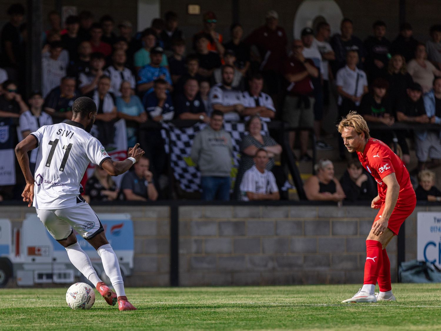 Two soccer players compete on a sunny pitch with a crowd of spectators watching in the background during a match.
