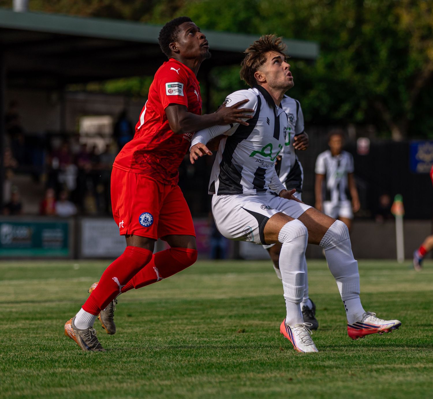 Two soccer players compete for the ball during a match, one in red and one in black and white striped kit.