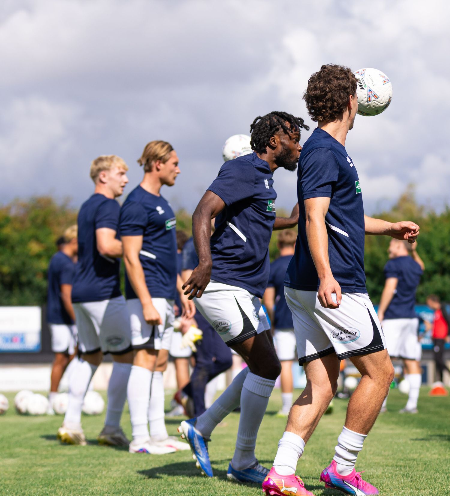 Soccer players in navy uniforms warm up on a green field, with one player heading a ball during practice.
