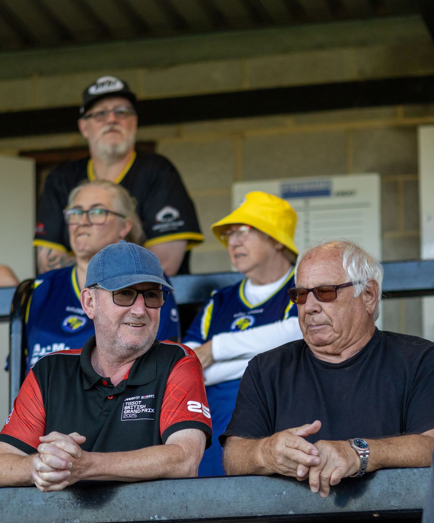 Football supporters of various ages watch a match from a stadium stand on a bright sunny afternoon.