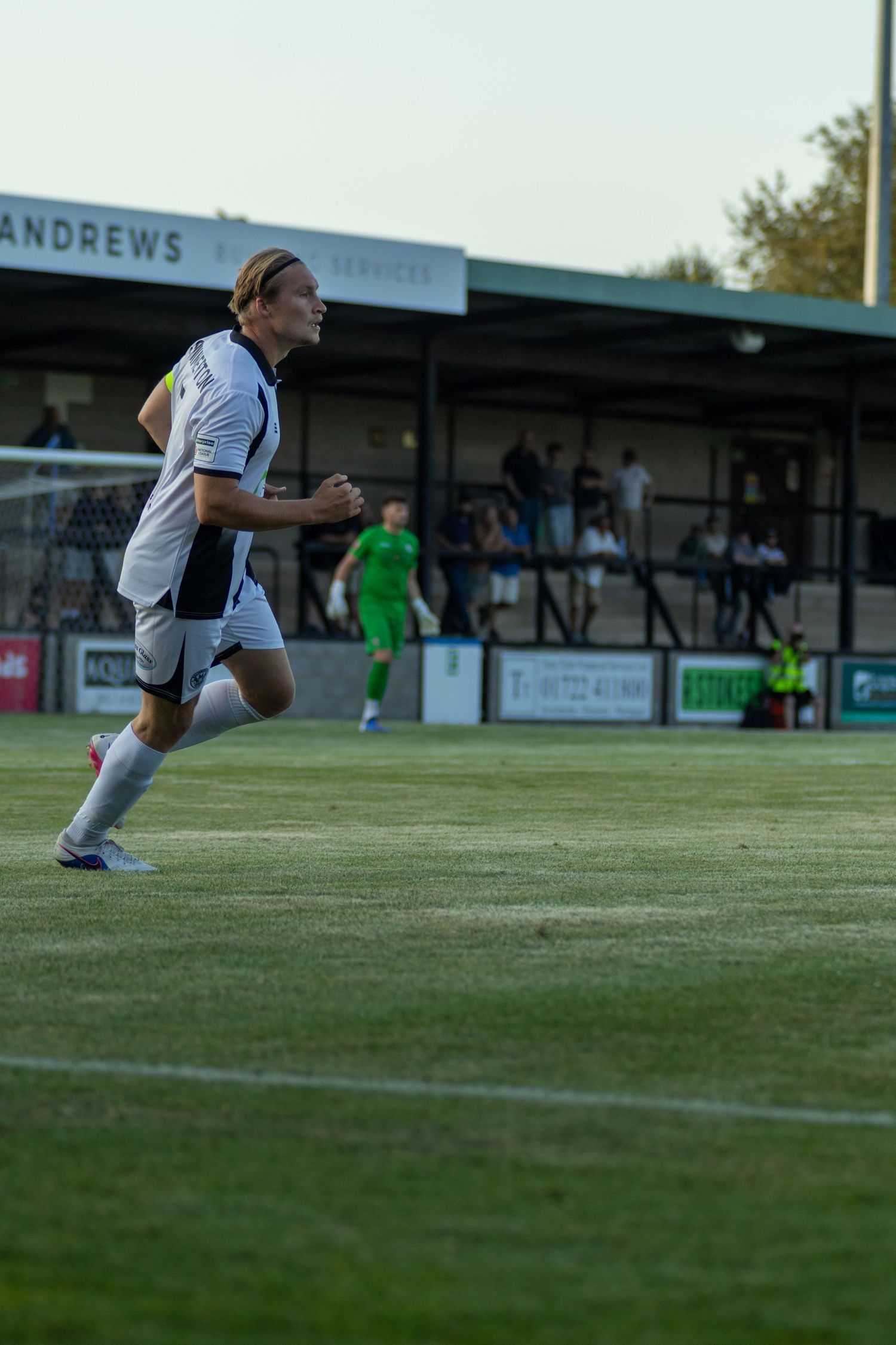 Soccer player in white kit sprinting across grass pitch with stadium stand visible behind.