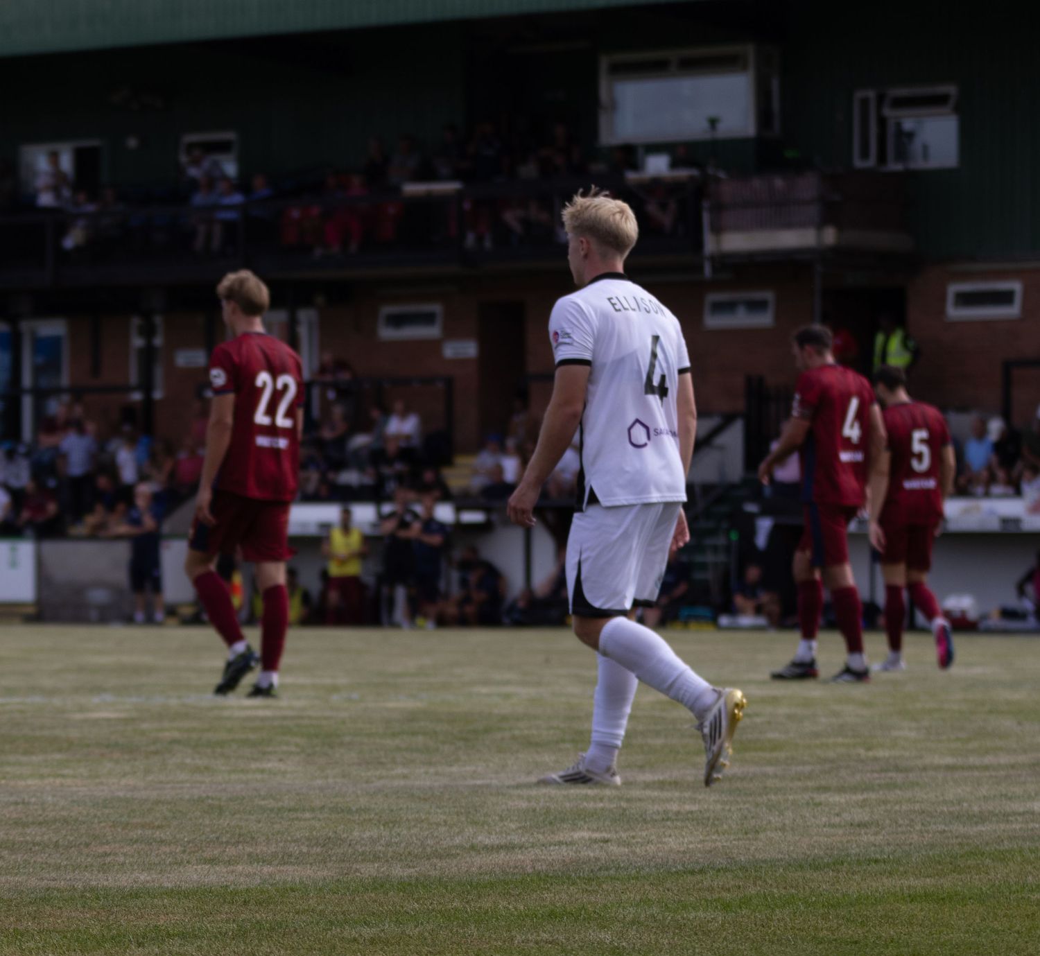 Football player wearing number 4 in white walks across the pitch during an outdoor football match.