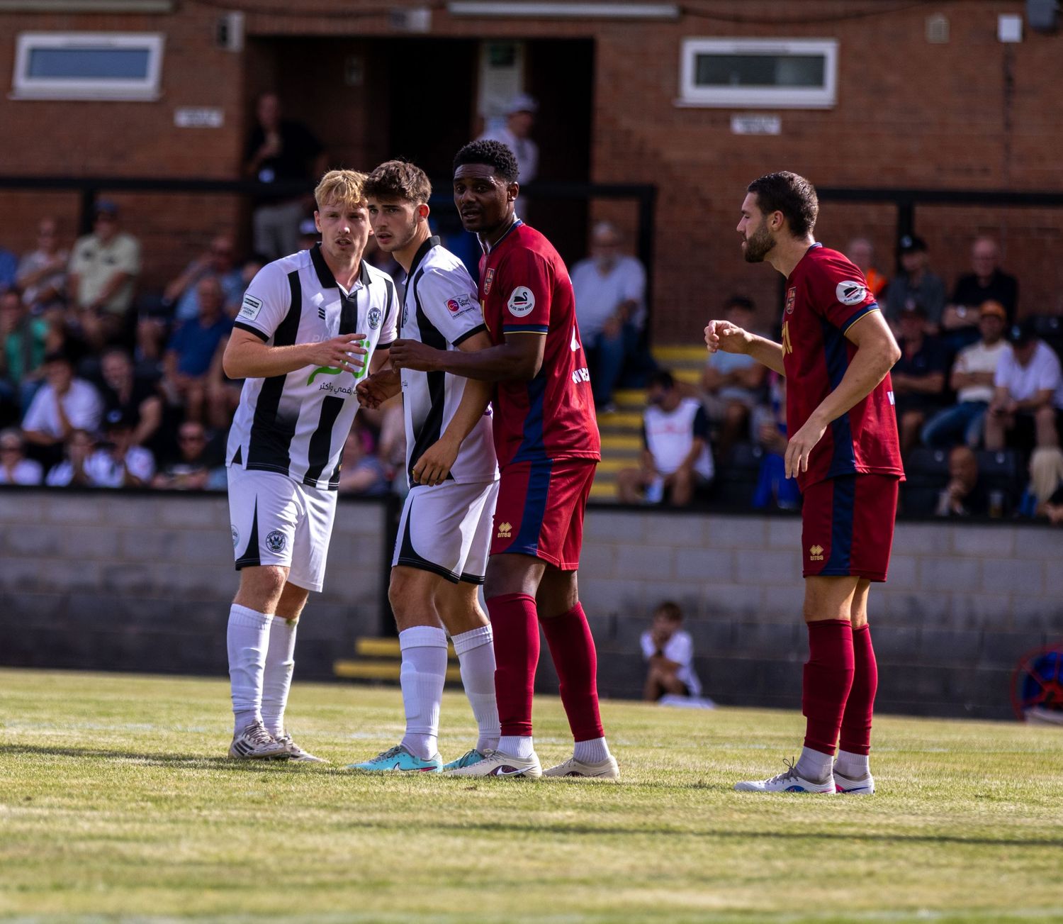 Four footballers in black-white and red kits face off during a heated moment in a non-league game.