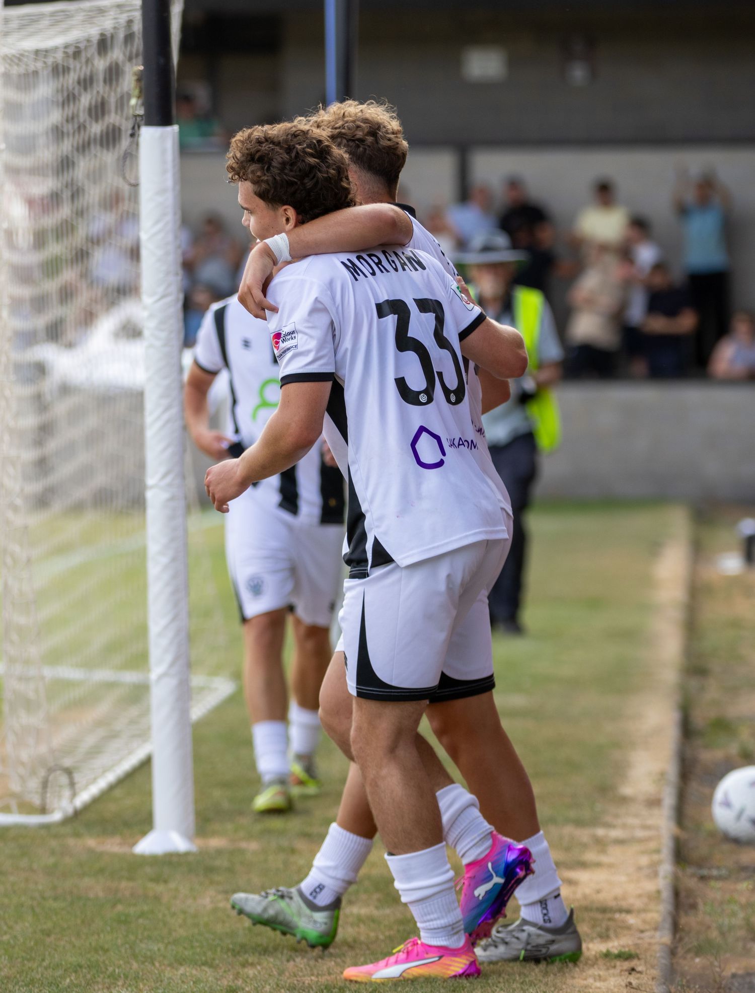 Two young footballers in white kits celebrate near the goalpost, with player number 33 Morgan hugging a teammate.