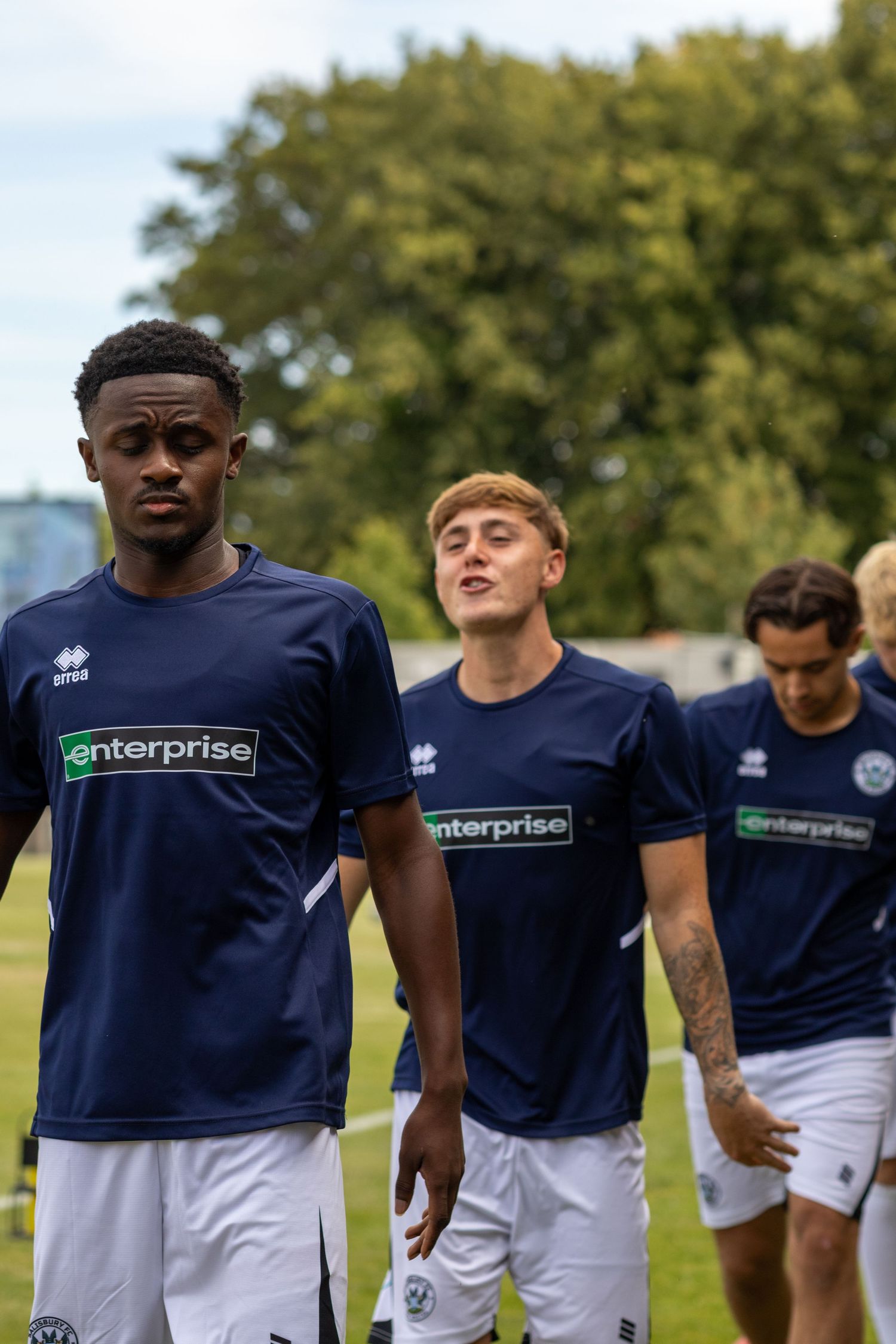 Amesbury FC players in navy Enterprise-sponsored shirts walk across the pitch during pre-match warmup.