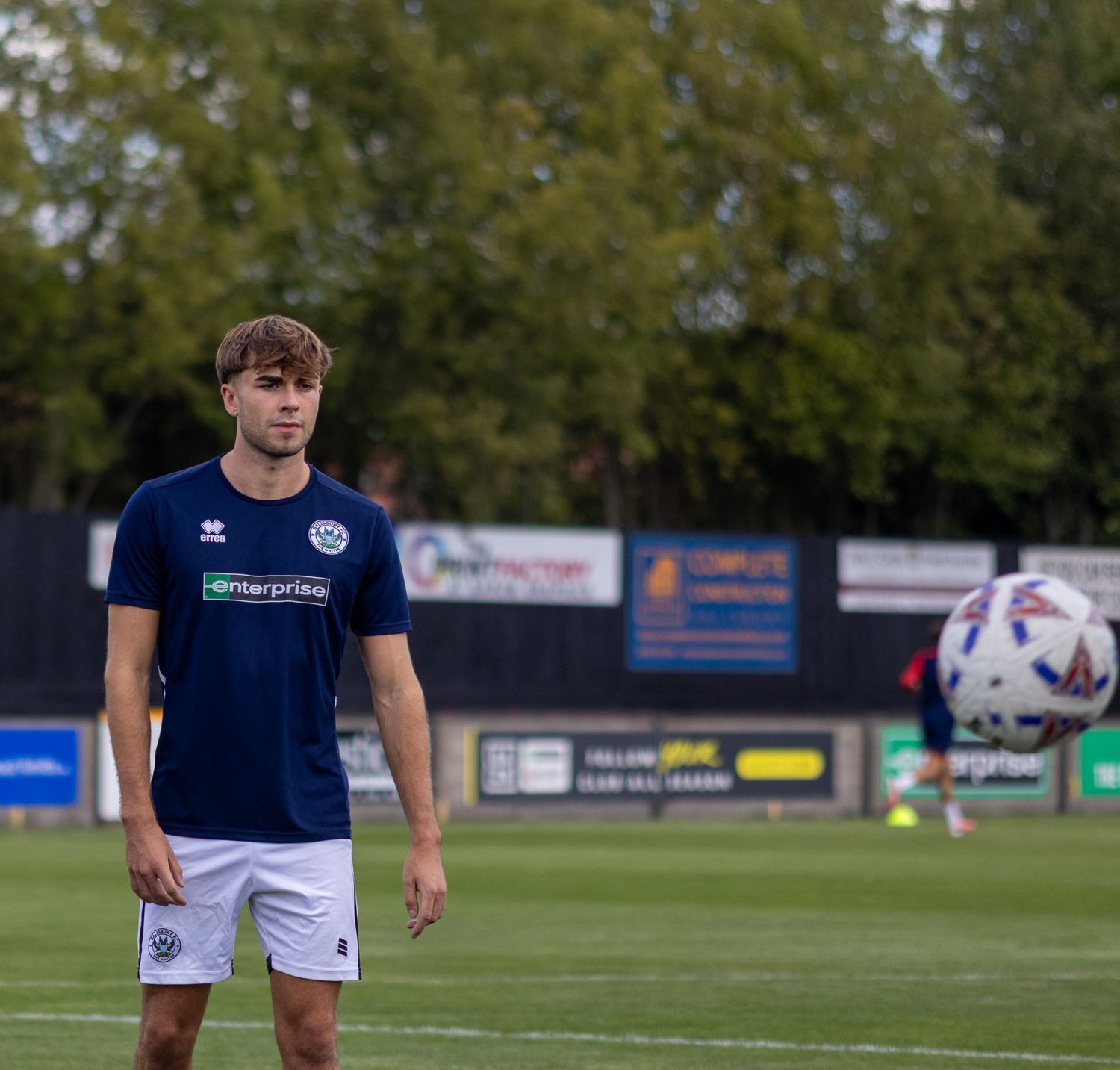 Young male footballer in navy enterprise-sponsored kit watches a ball during pre-match warmup on grass pitch.