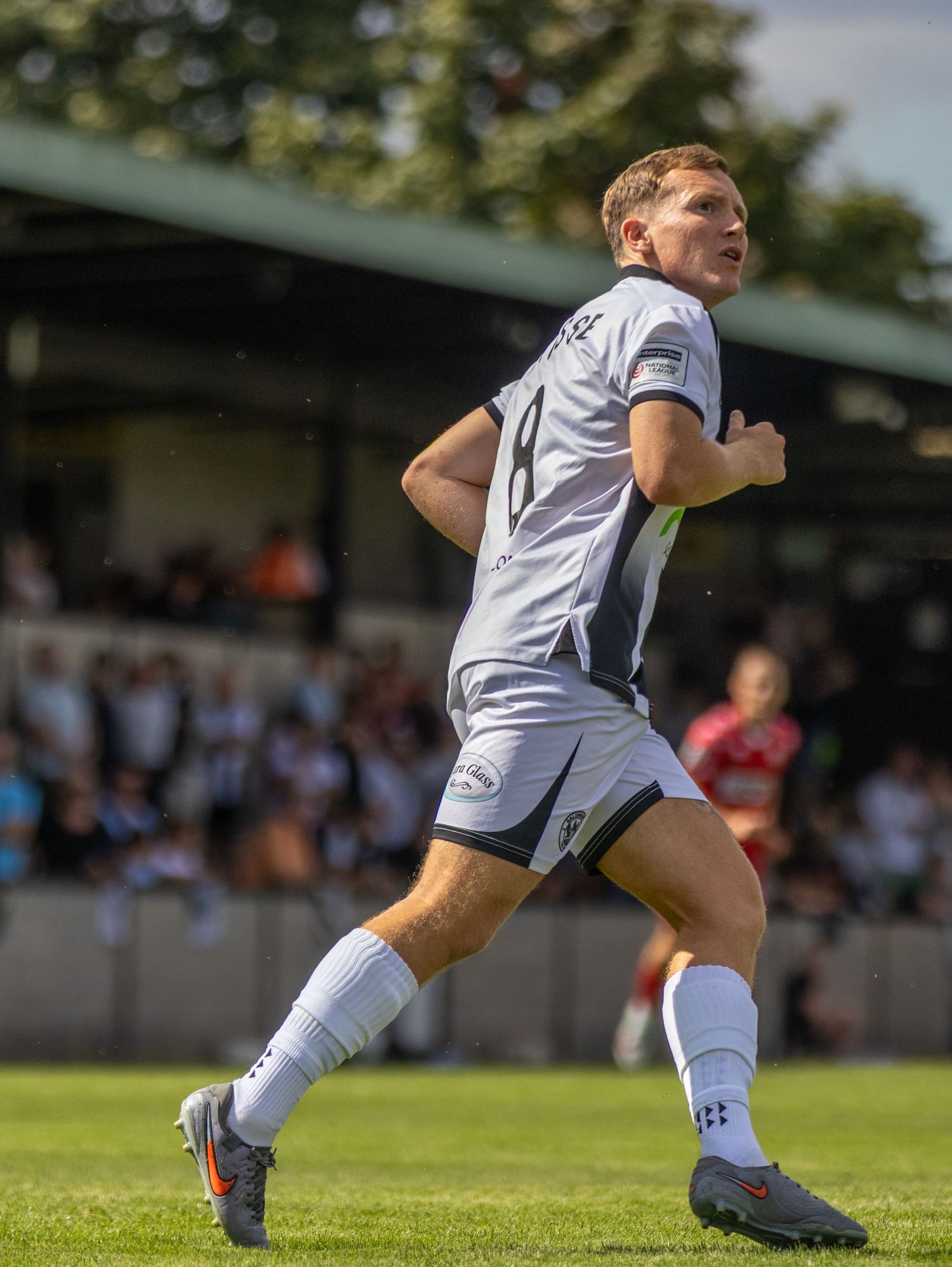 A male footballer in white kit sprints across green pitch with crowd visible in the background.