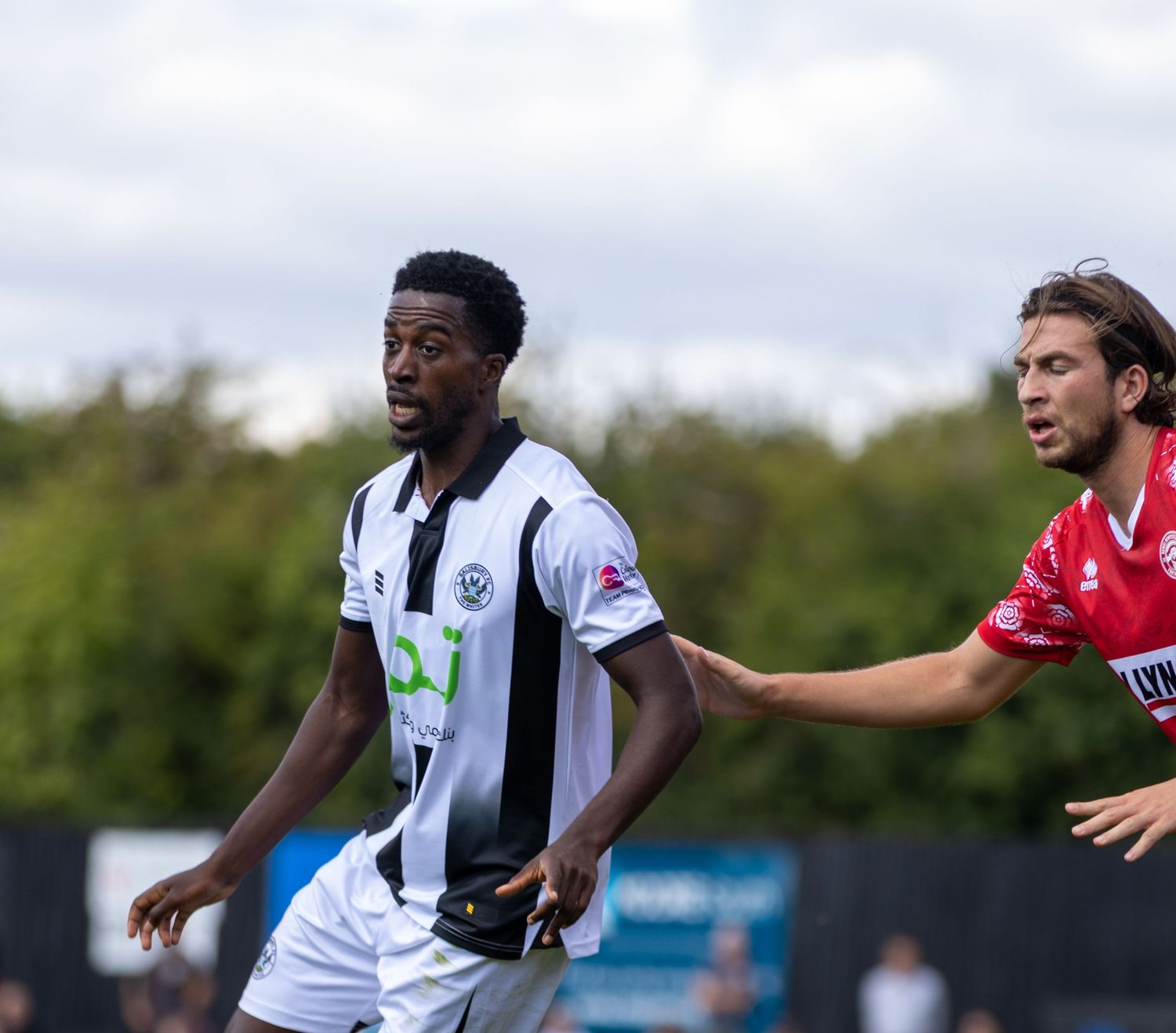Black and white kit footballer contests possession with a red-kitted opponent during an outdoor football match.