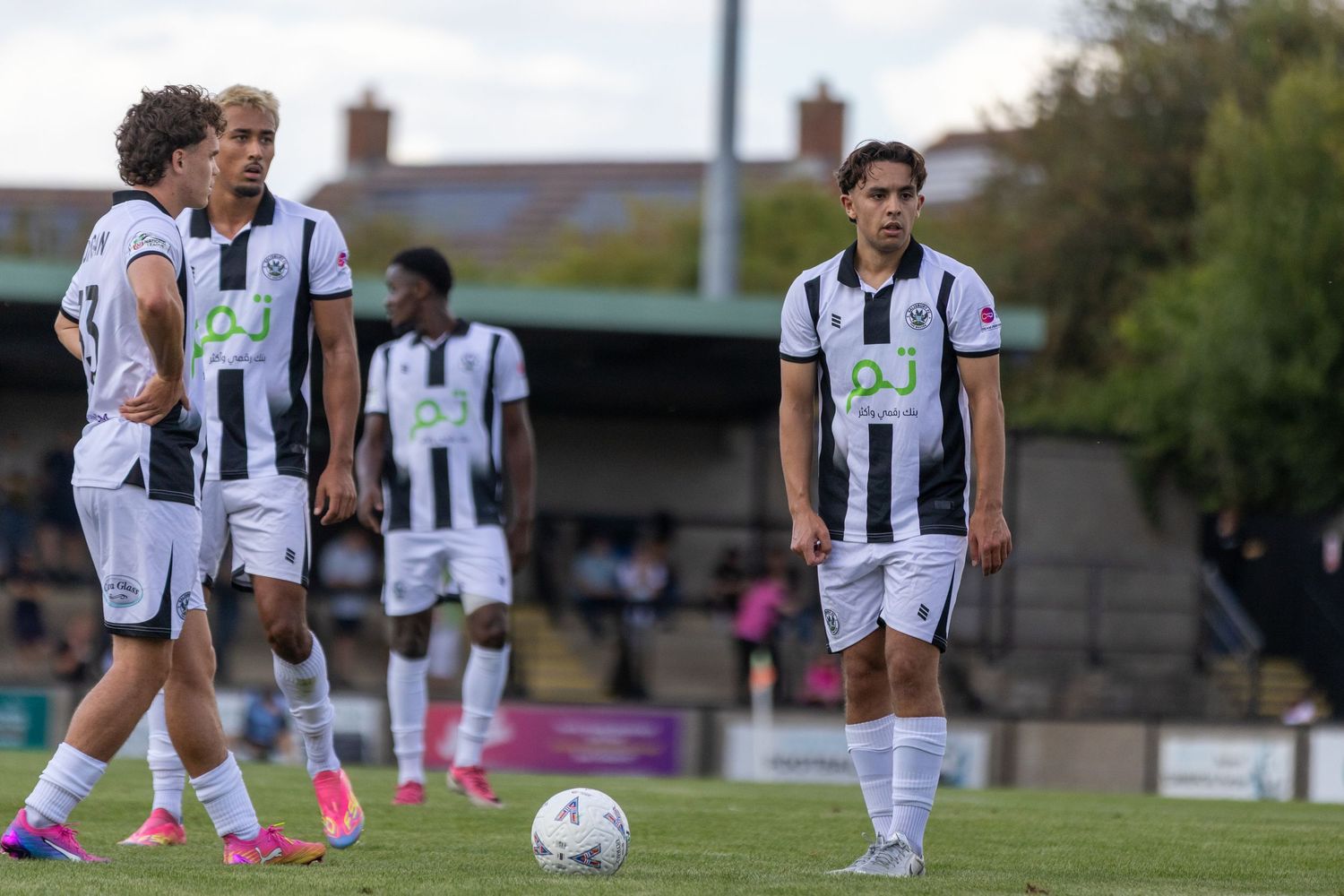 Black and white football players prepare for a free kick on a grass pitch during a match.