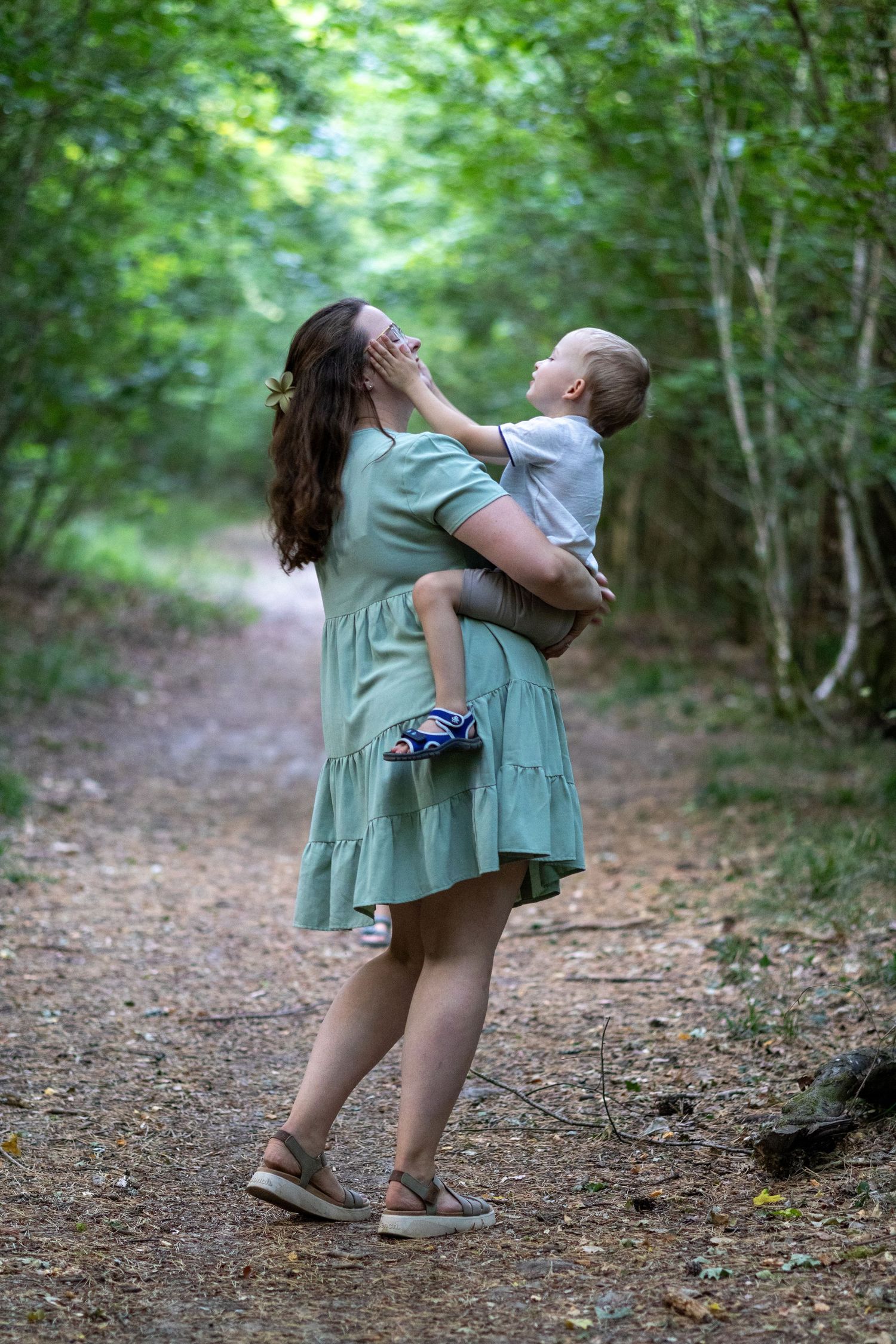 Smiling mother in mint dress carrying toddler son on a green forest path.